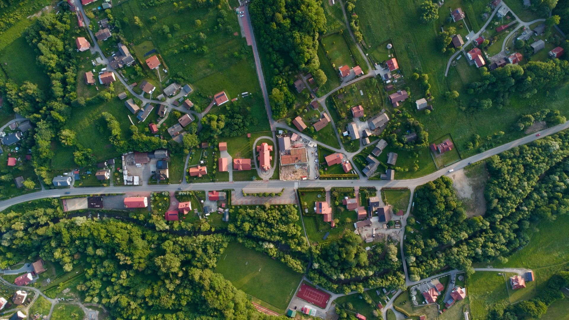 Aerial view of a village with red-roofed buildings, a central road, and surrounding green trees and fields.