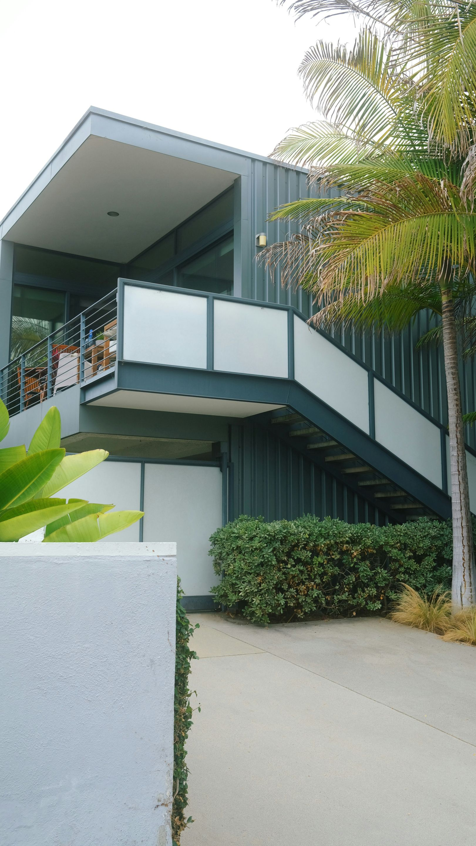Modern dark gray house with a prominent exterior staircase, glass balcony, and palm trees in a sunny, paved driveway.