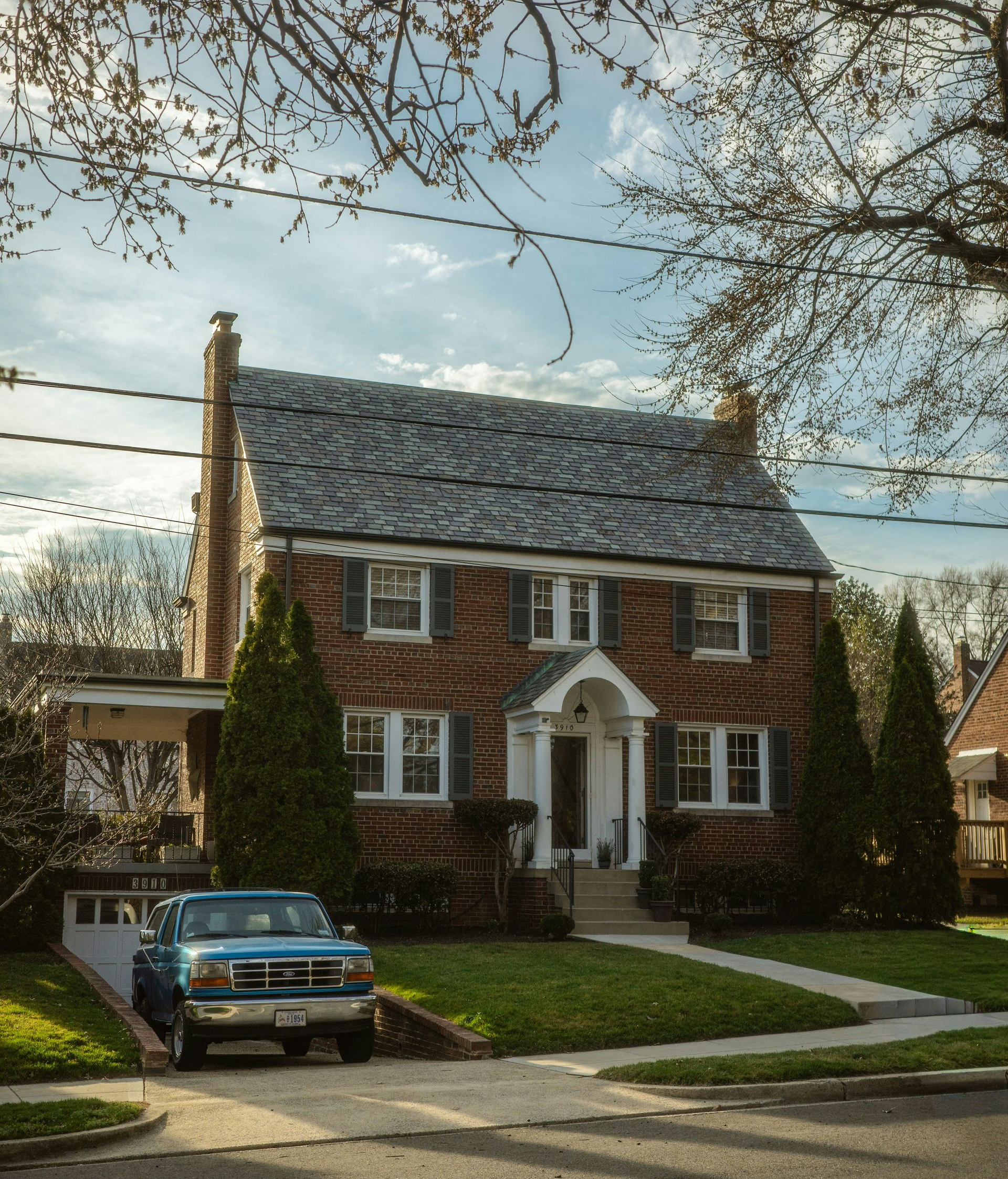 A brick two-story house with a grey shingled roof, a front porch, and a blue pickup truck parked in the driveway.