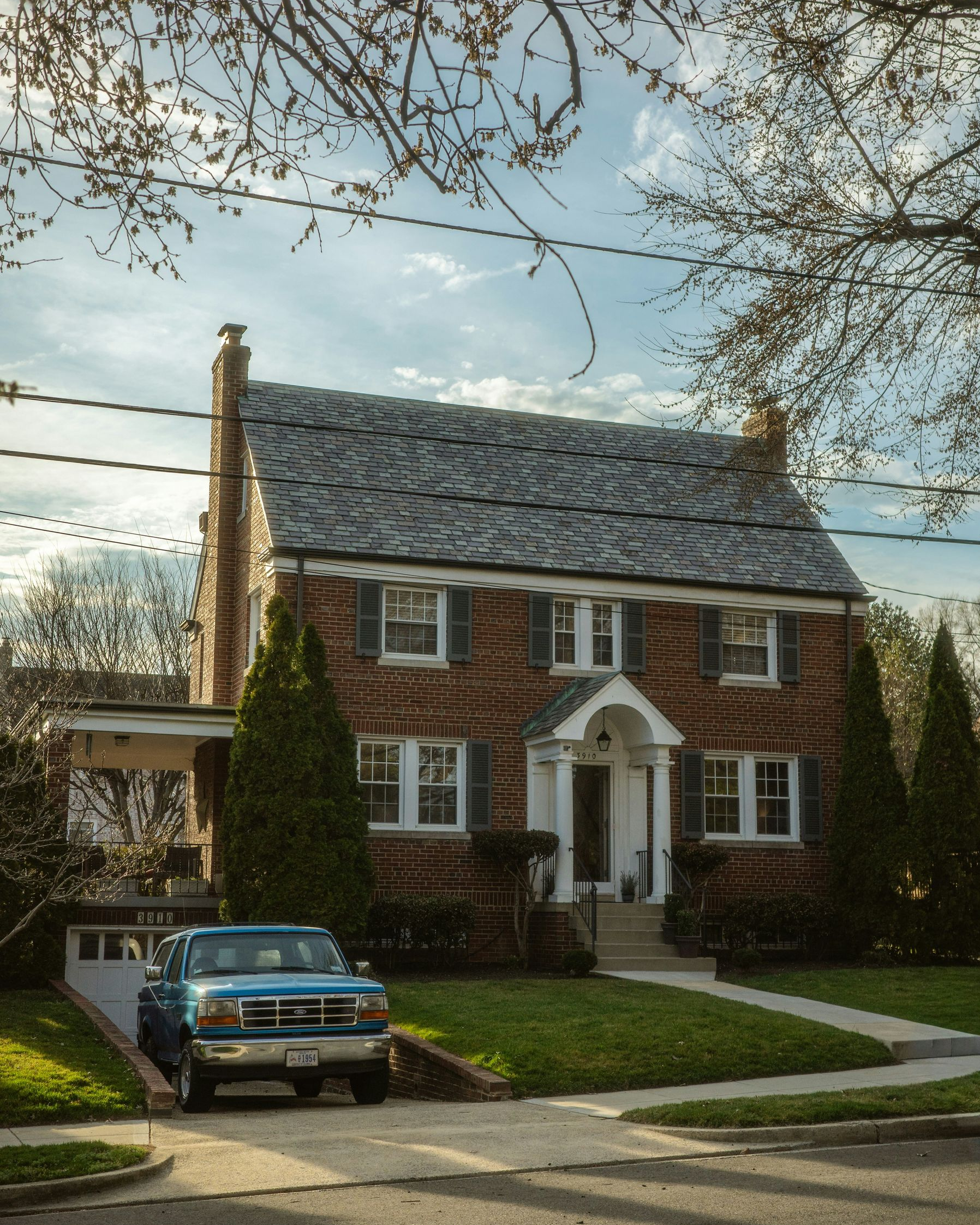 A brick two-story house with a grey shingled roof, a front porch, and a blue pickup truck parked in the driveway.