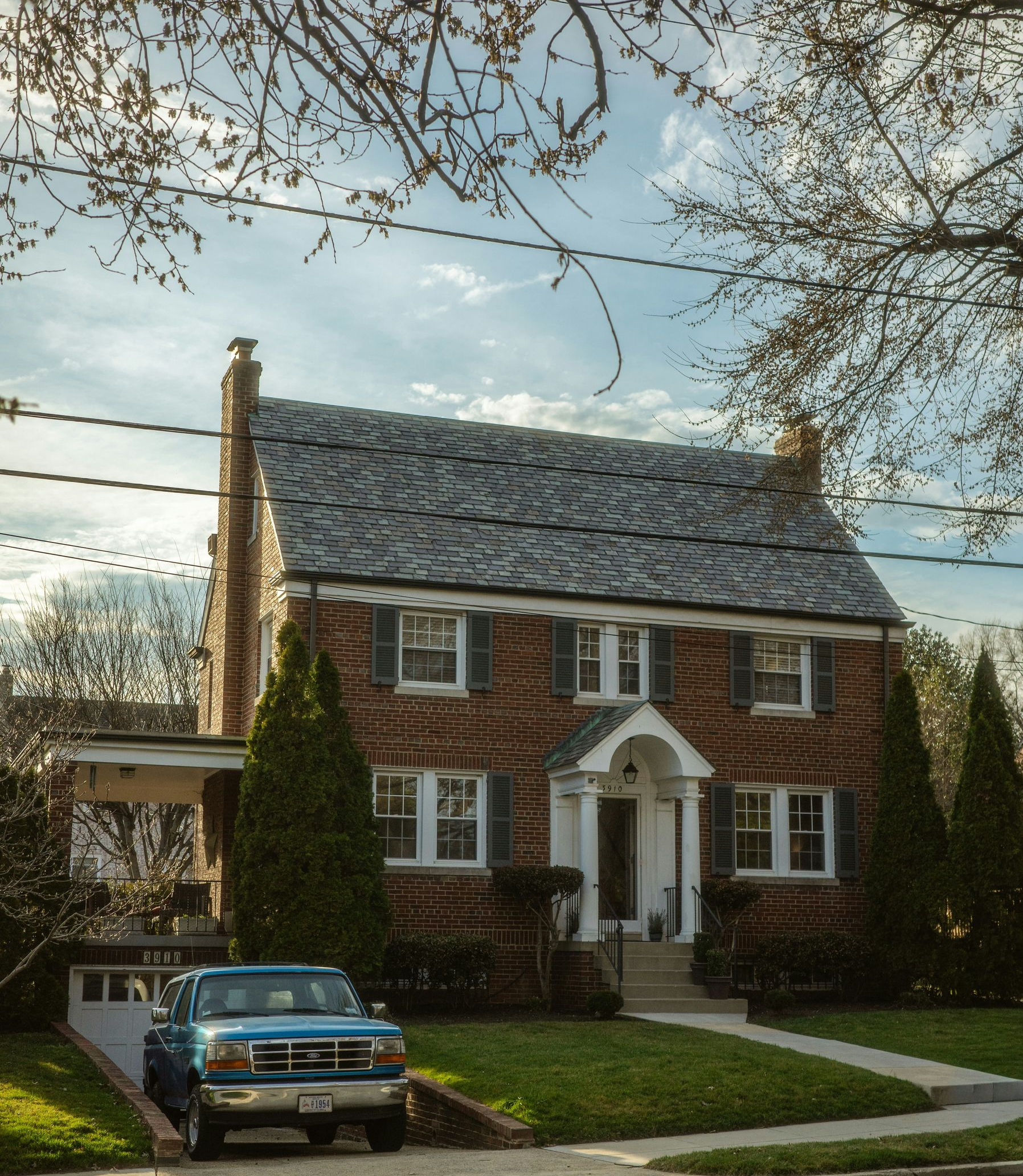 A brick two-story house with a grey shingled roof, a front porch, and a blue pickup truck parked in the driveway.