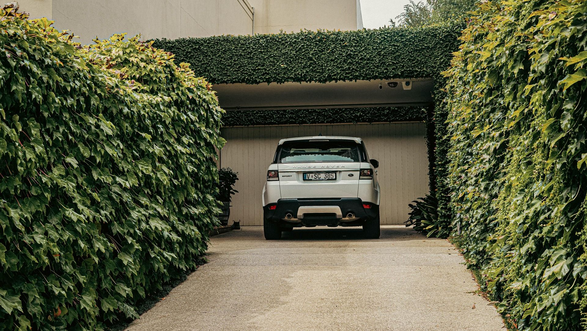 A white SUV parked in a gravel driveway framed by lush green ivy walls leading toward a covered carport.