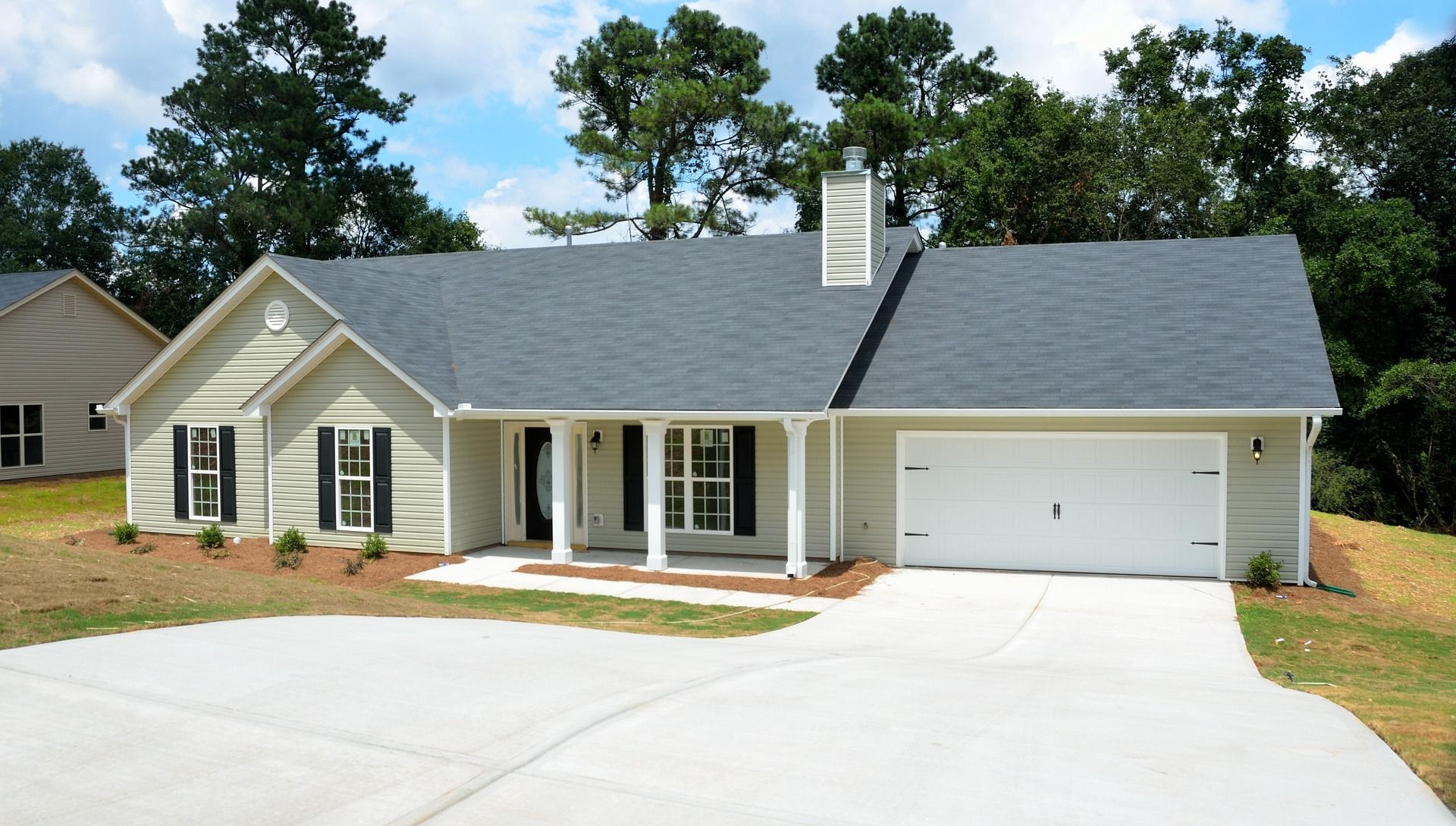 A light-colored single-story house with a white garage, dark shingled roof, and a front porch, set among green trees.