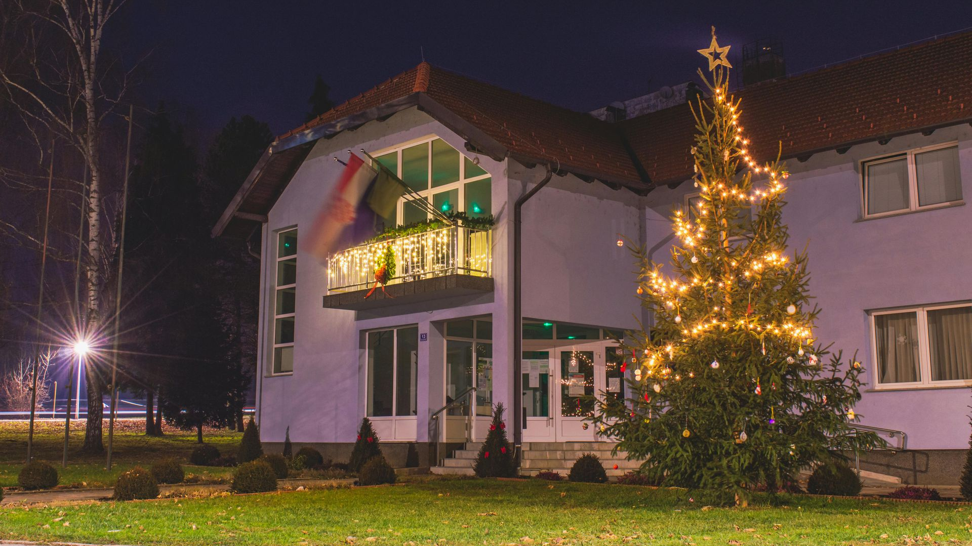 Building lit at night with Christmas tree lights in front. Flags on balcony.