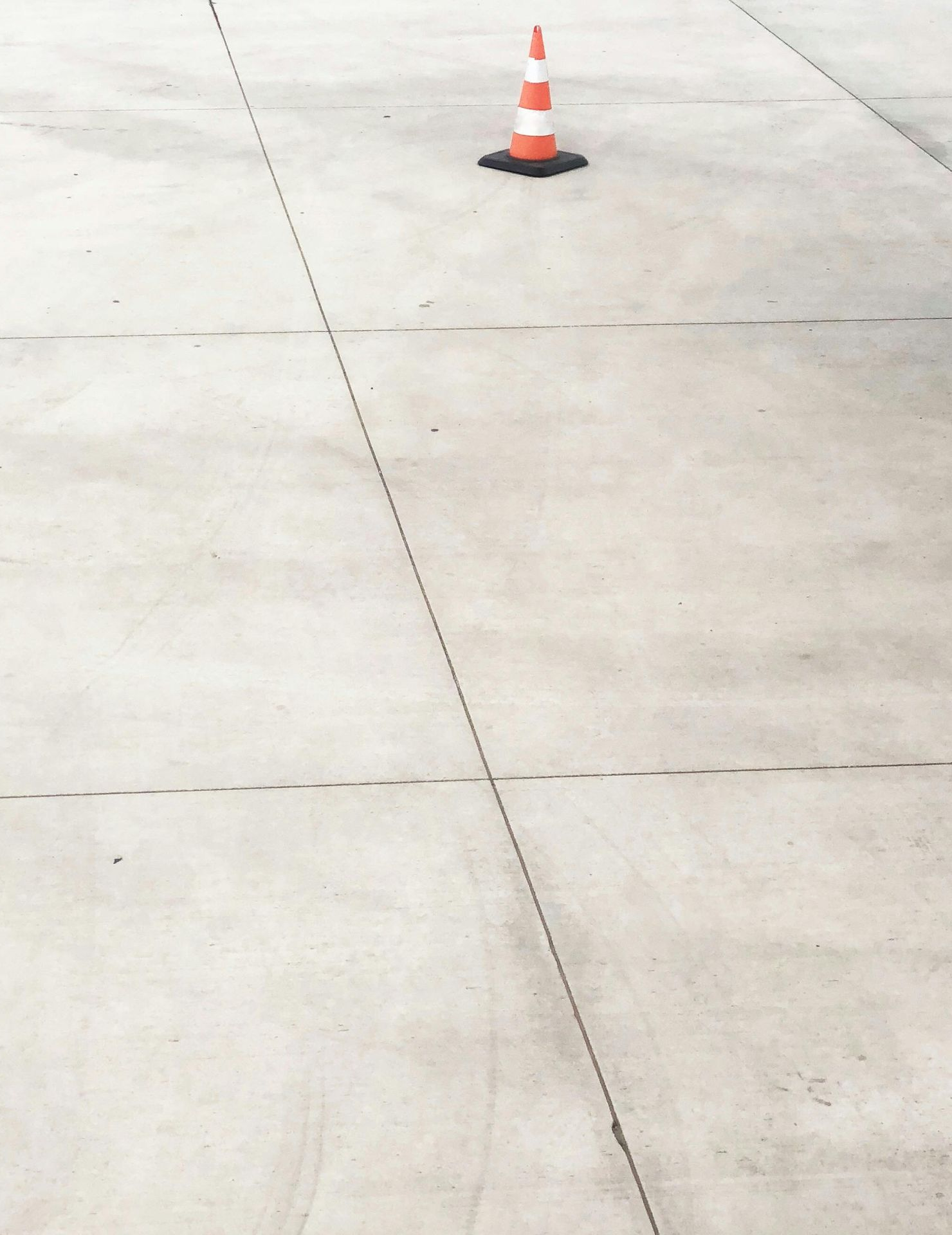 A single orange and white traffic cone sits alone on a wide expanse of light gray concrete pavement.