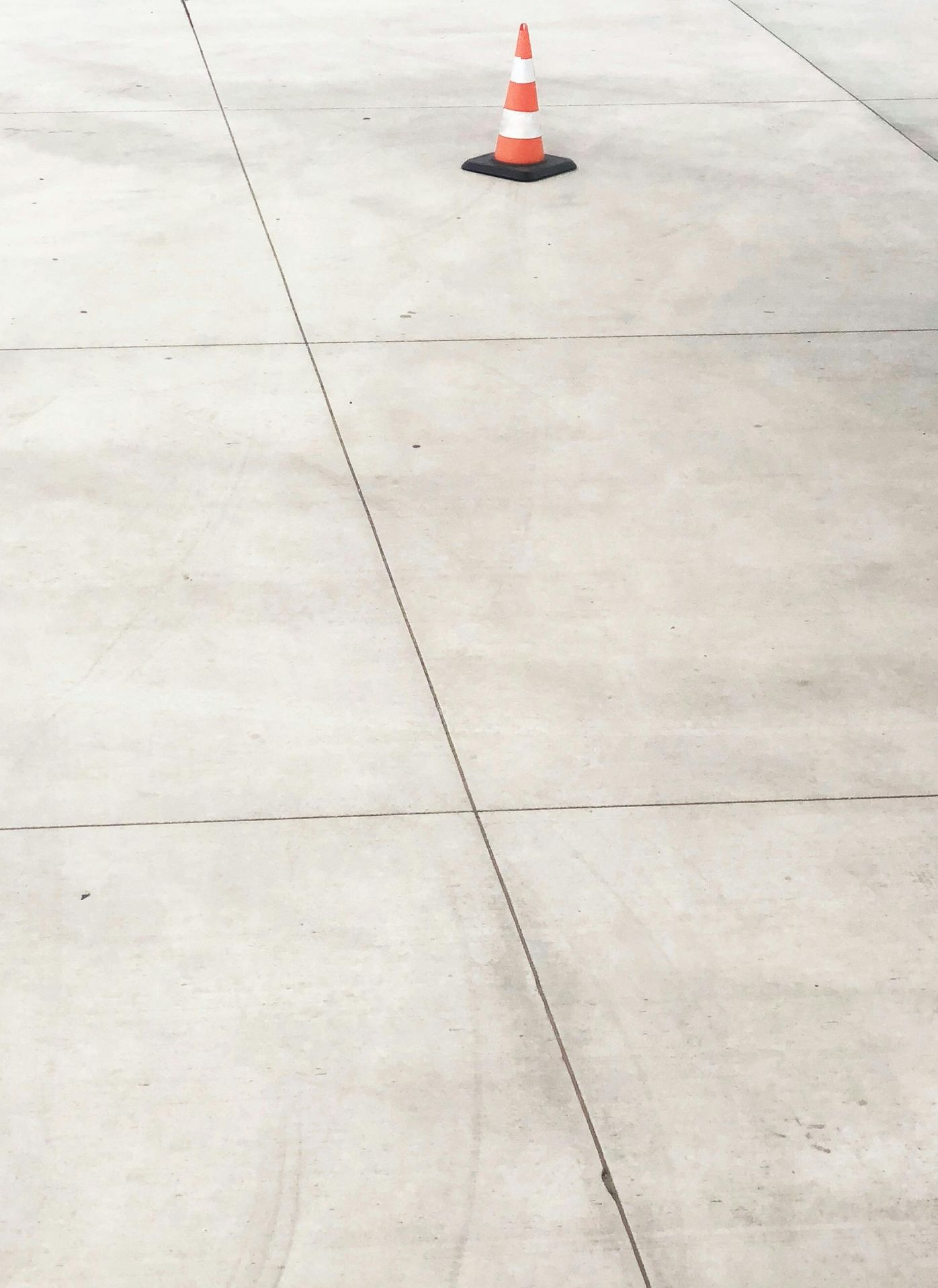 A single orange and white traffic cone sits alone on a wide expanse of light gray concrete pavement.