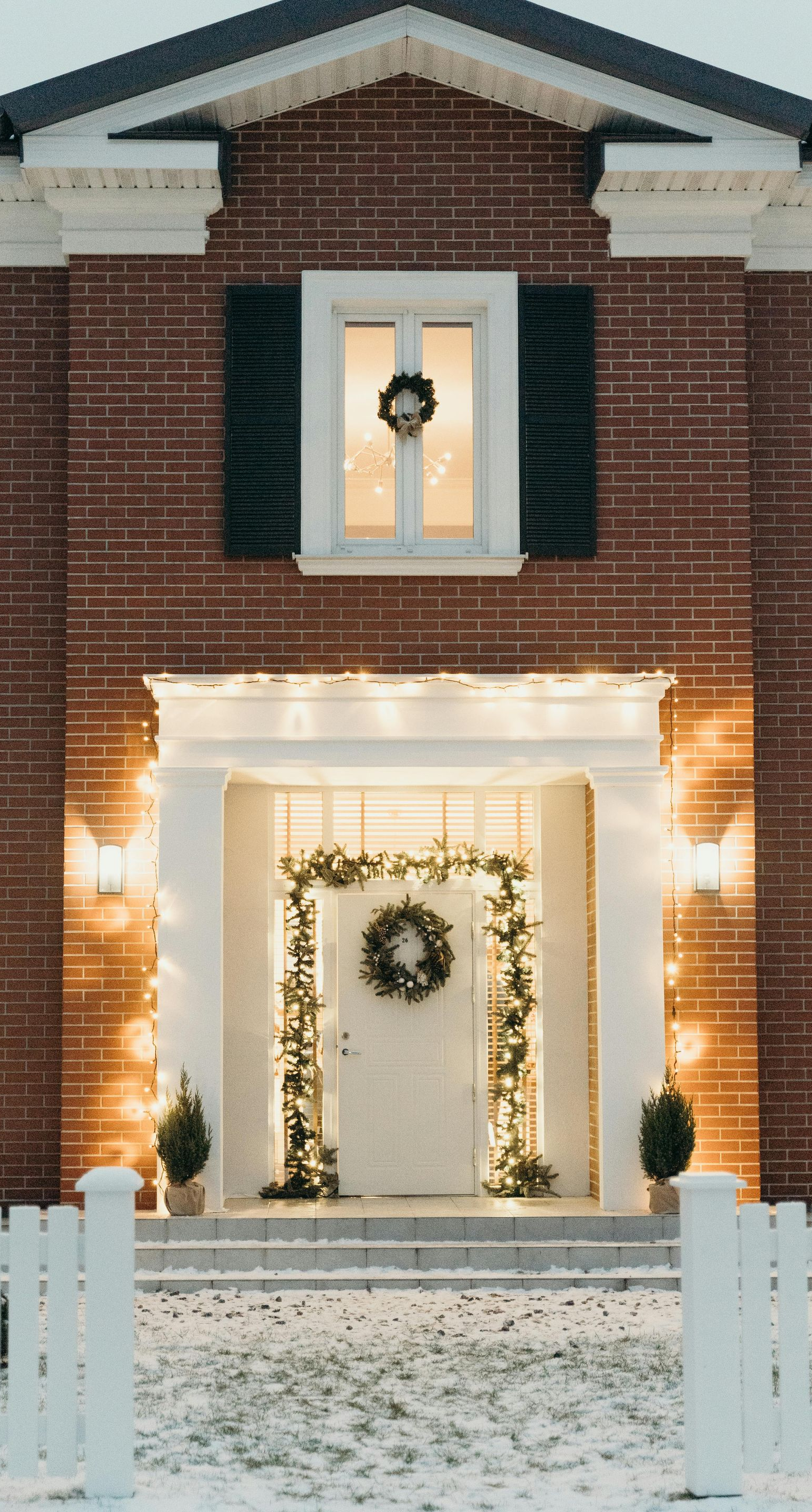 Festive brick home entrance with snow, lit garlands, wreaths, and white fence.