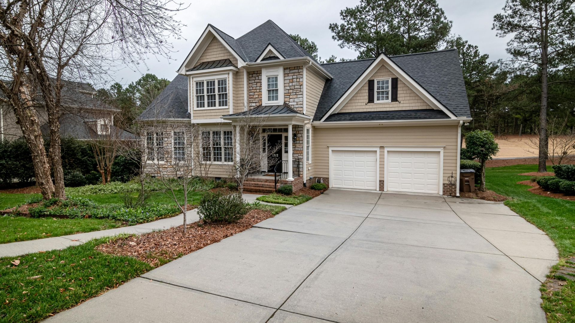Tan two-story house with black roof and two-car garage; paved driveway leads to the house, with green grass and trees surrounding.