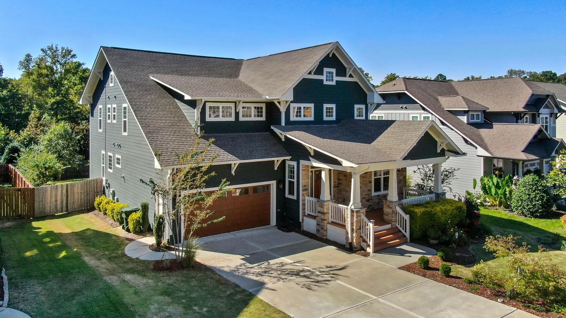 A multi-story, dark green home with a brown garage and stone accents, viewed from an elevated angle on a sunny day.