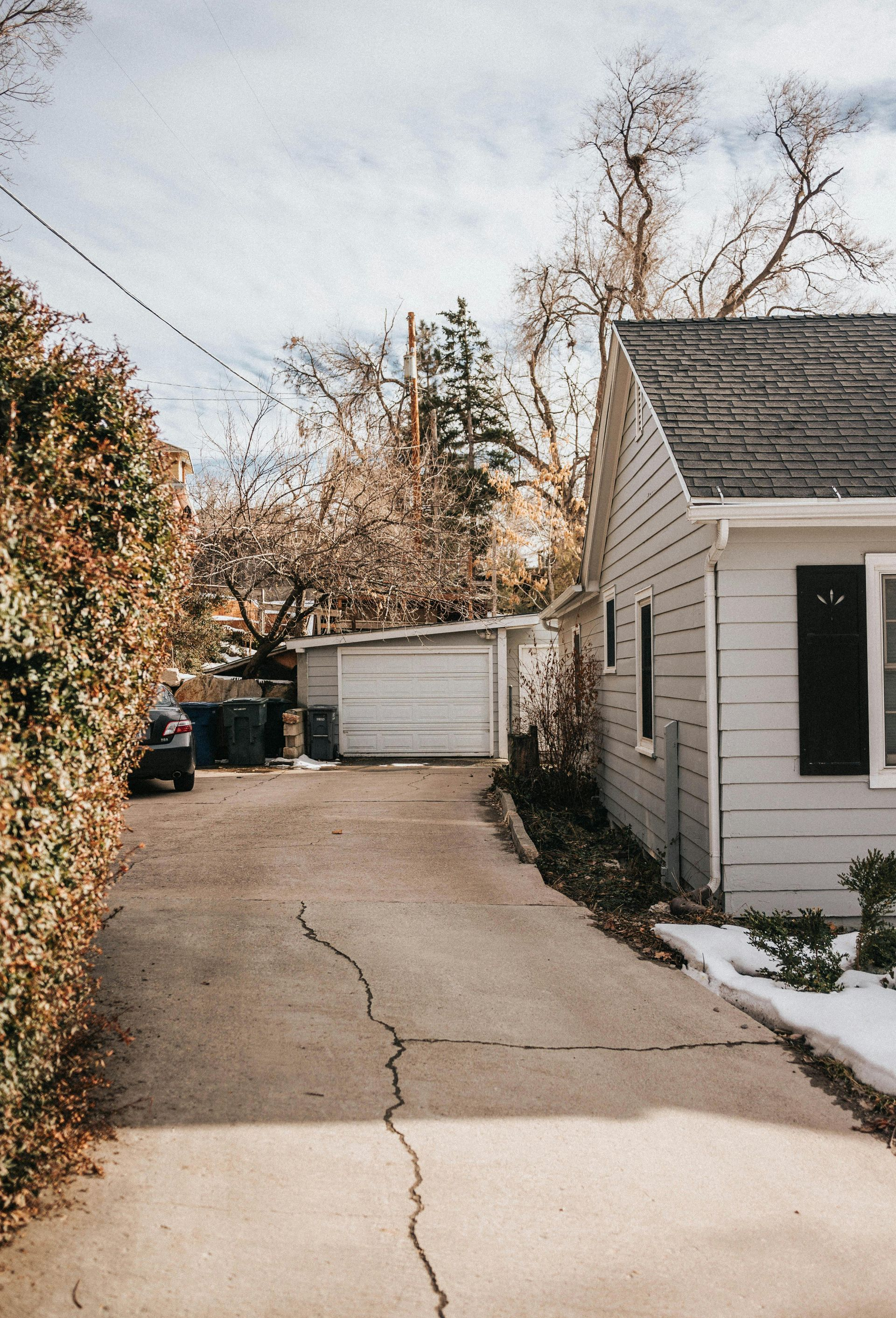 A light-colored house with a dark roof sits beside a long driveway leading to a white detached garage, with trees above.