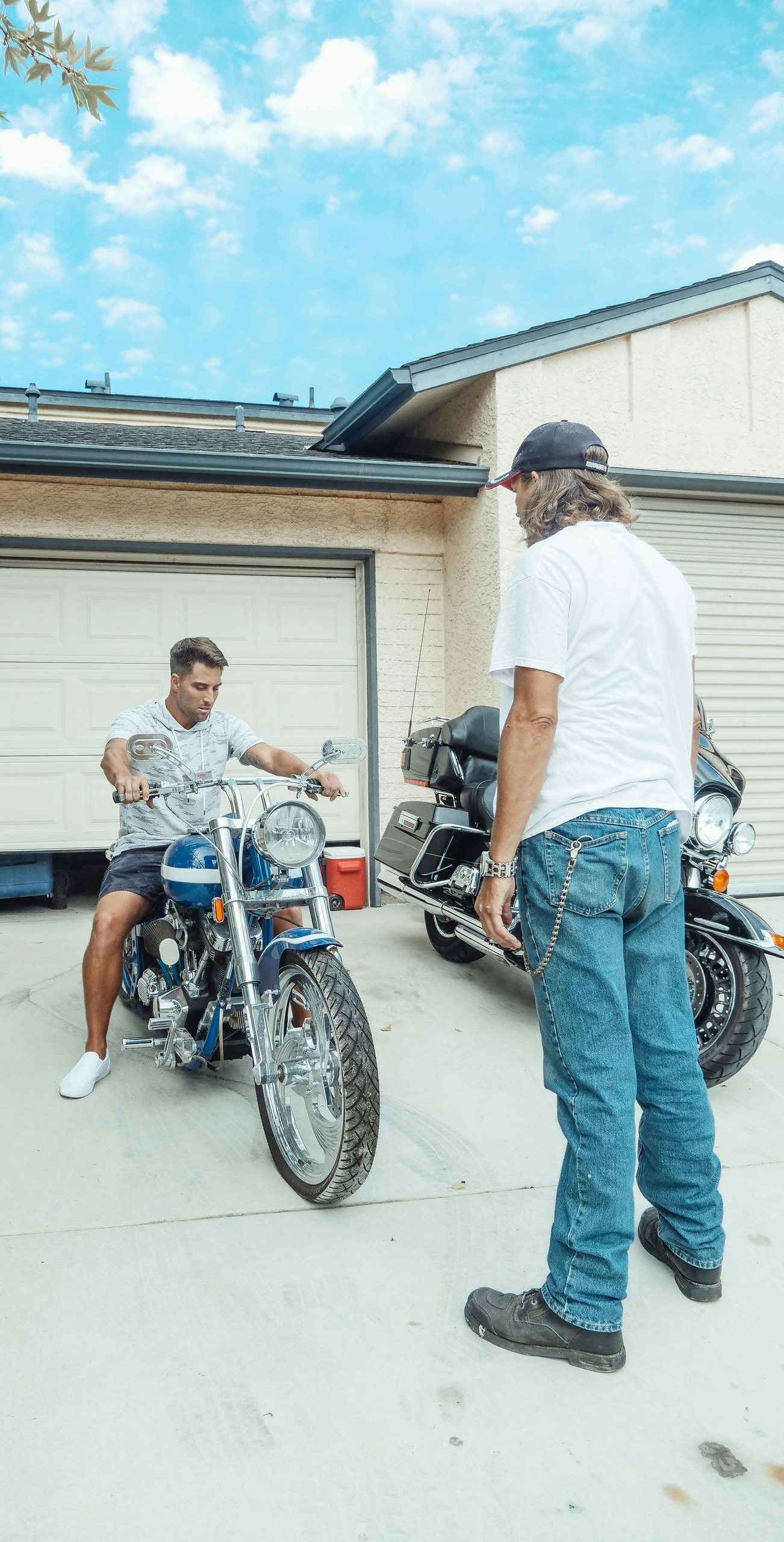 Two men near motorcycles outside a garage. One sits on a blue chopper, the other stands beside a black touring bike. Blue sky.