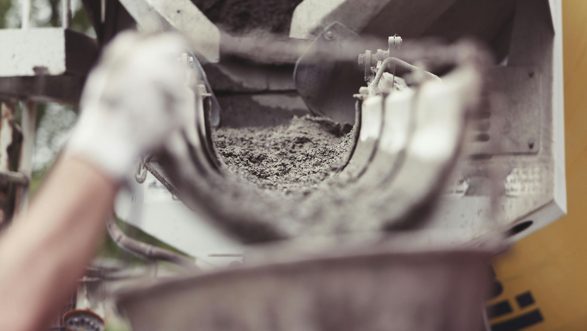A person's gloved hand pouring concrete mixture into a machine on a construction site.