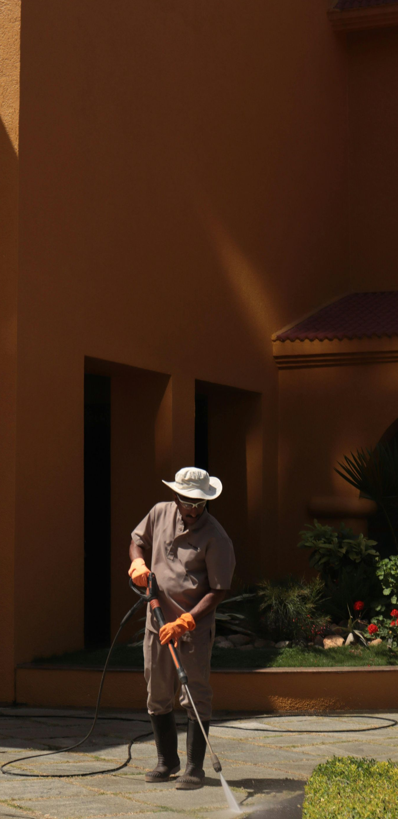 Person in hat and gloves pressure washing a concrete patio next to a building.