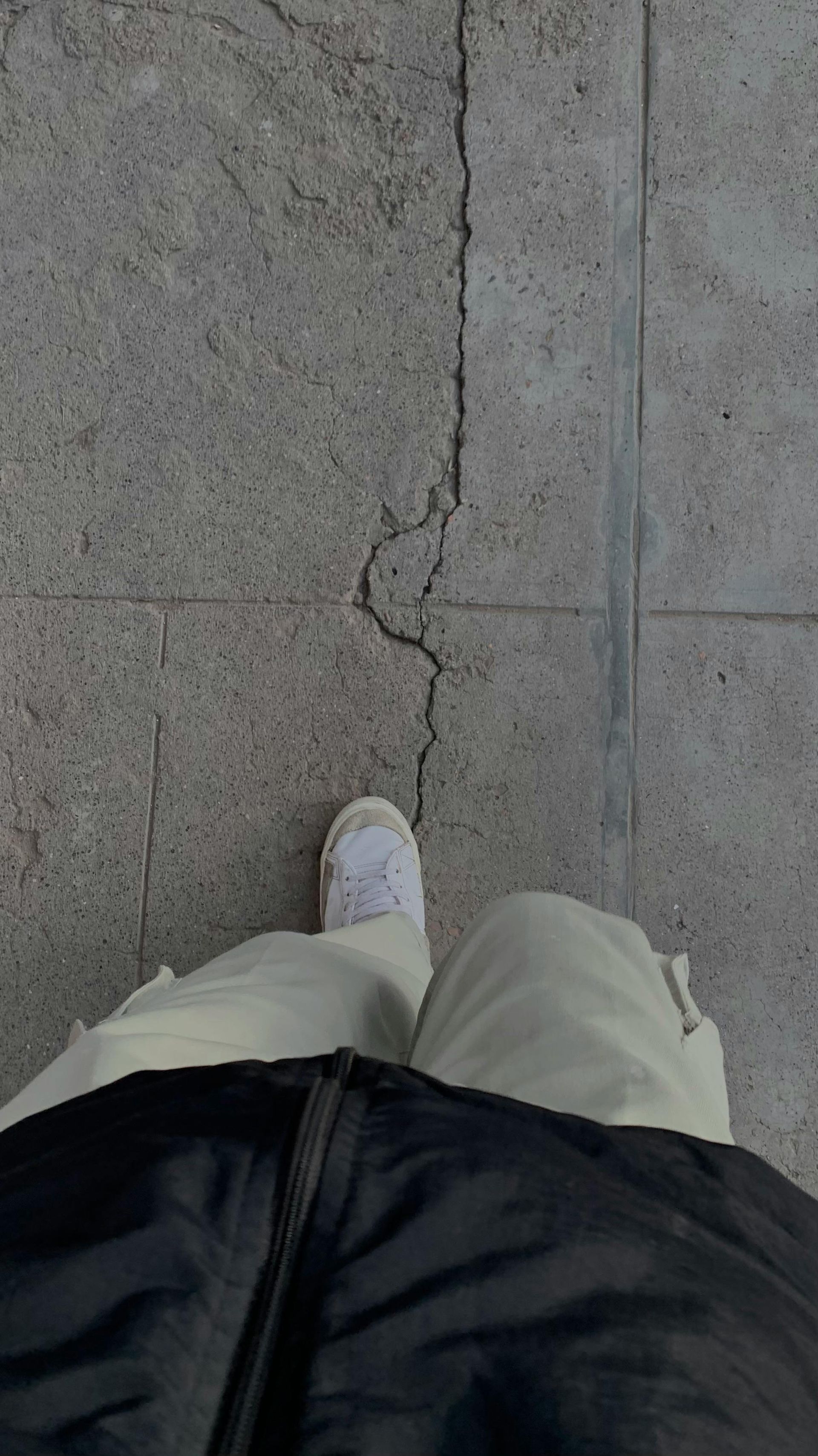 Person's feet on cracked pavement. White sneakers, beige pants, and black jacket visible.