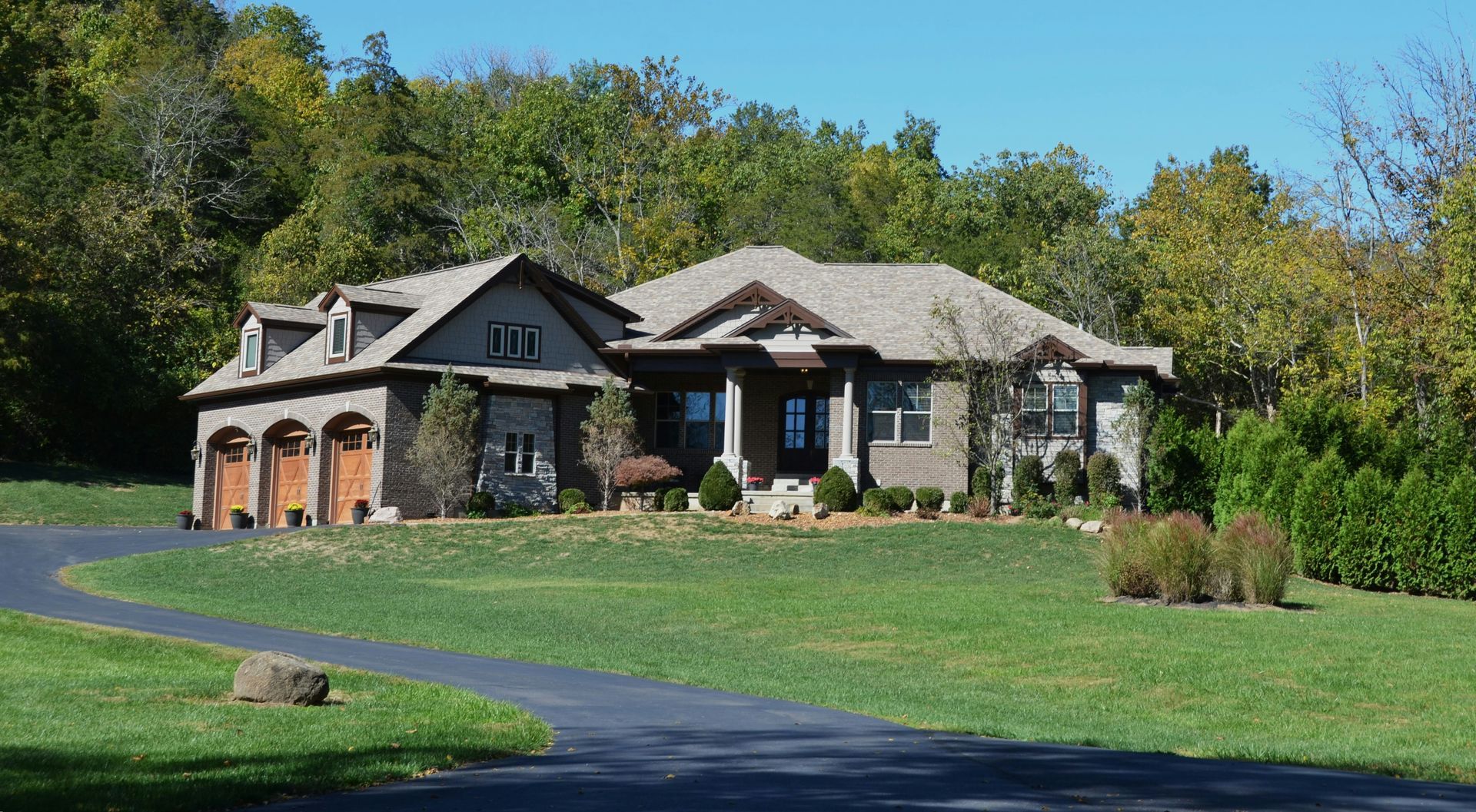 House with attached garage, stone and wood exterior, set on green lawn with long driveway, surrounded by trees.