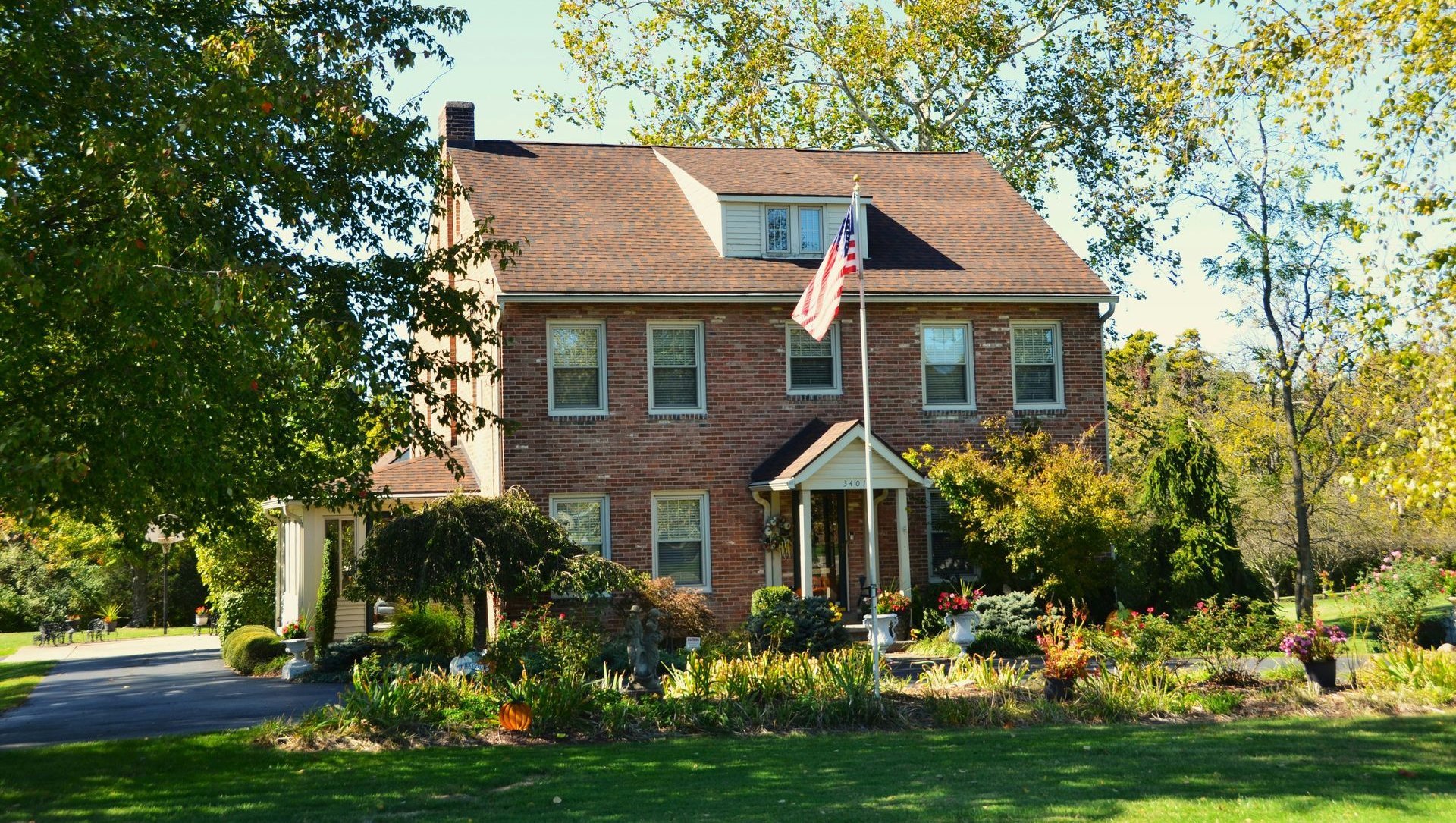 A two-story brick house with a brown roof and dormer window, surrounded by trees, a garden, and an American flag.