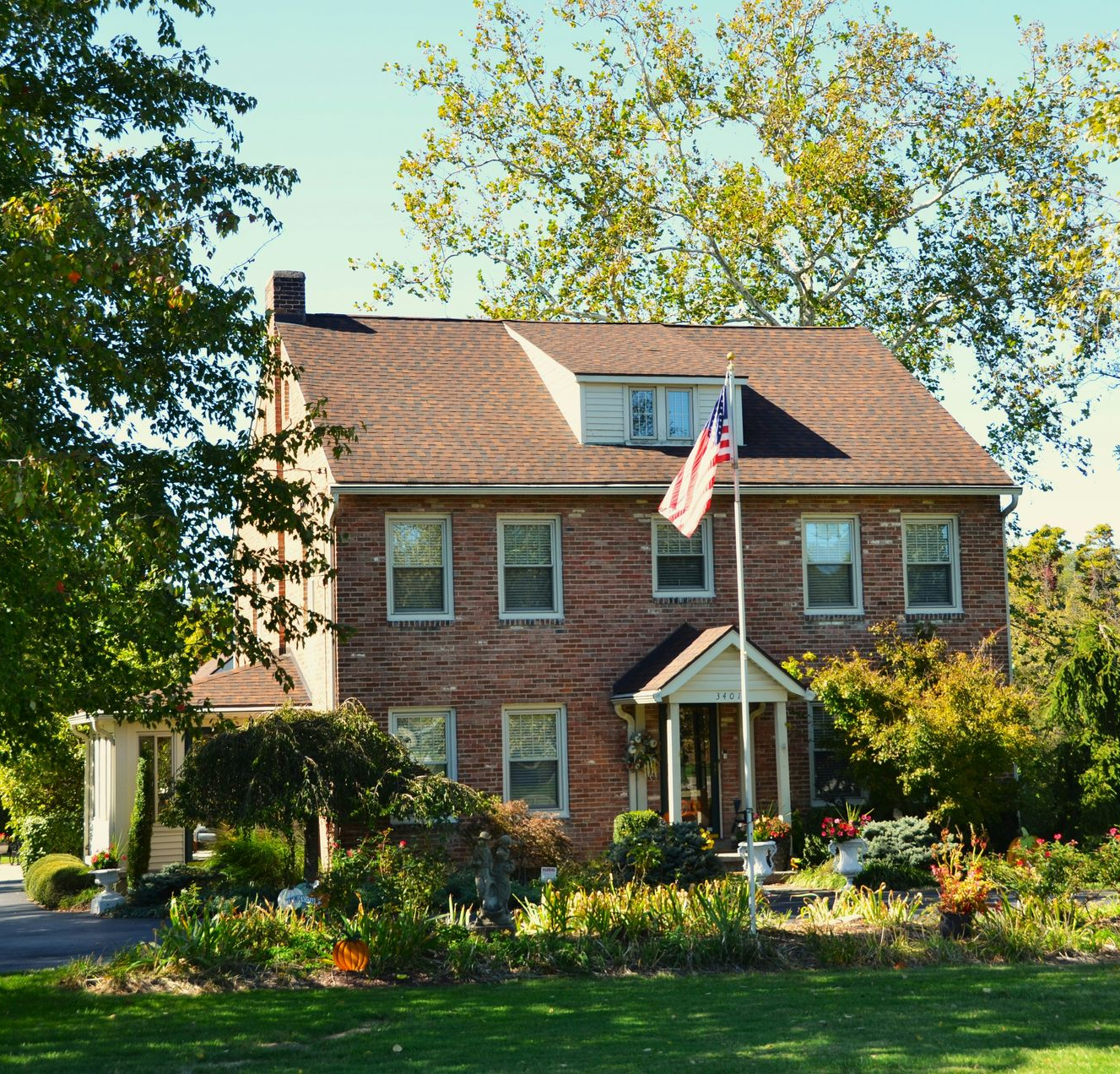Brick house with American flag, surrounded by trees and a garden on a sunny day.