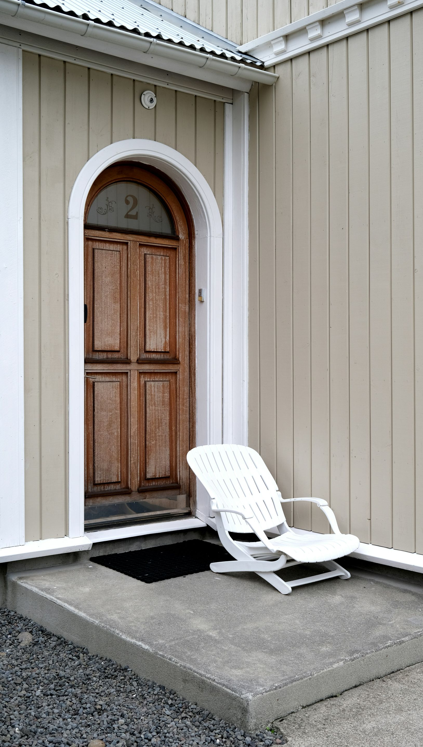 Wooden door in an arched doorway with a white chair on the concrete step. Light tan building exterior.