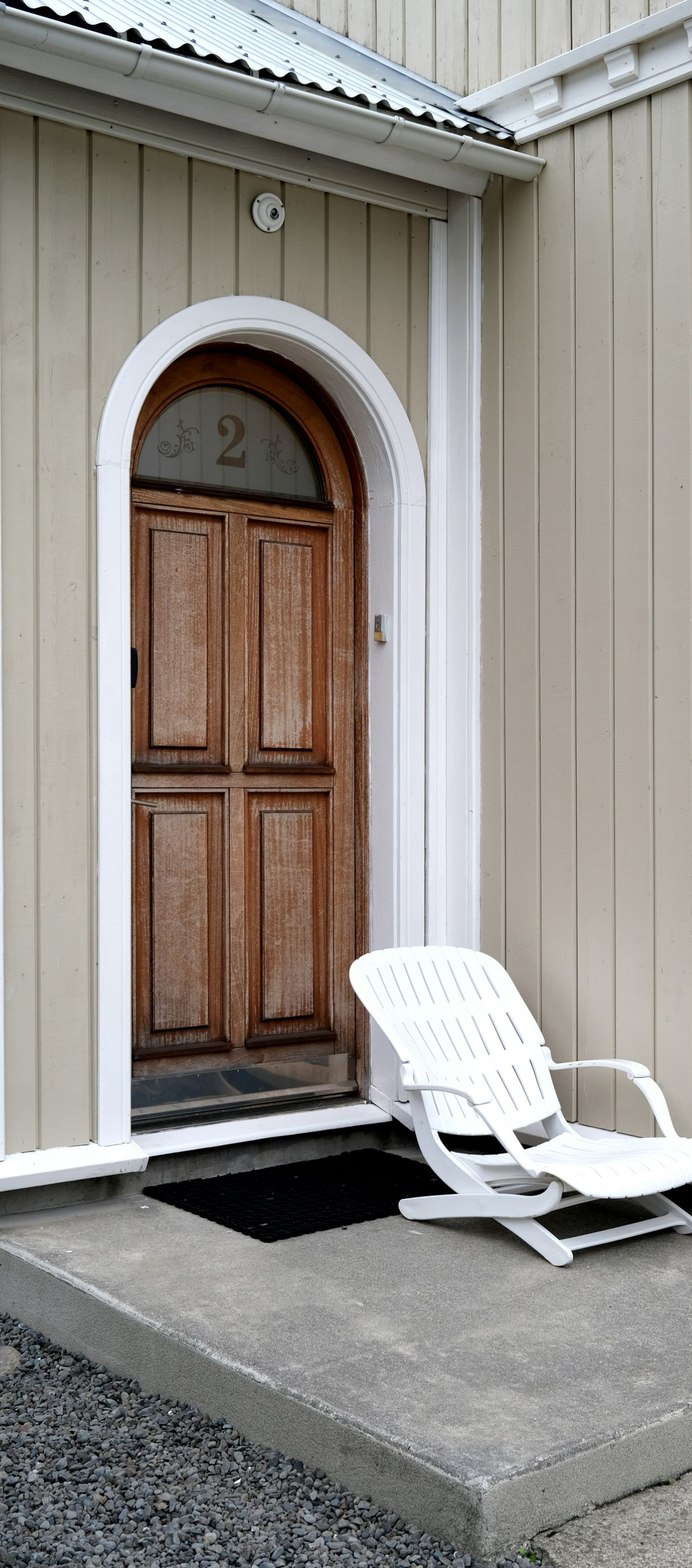 Wooden door in an arched doorway with a white chair on the concrete step. Light tan building exterior.