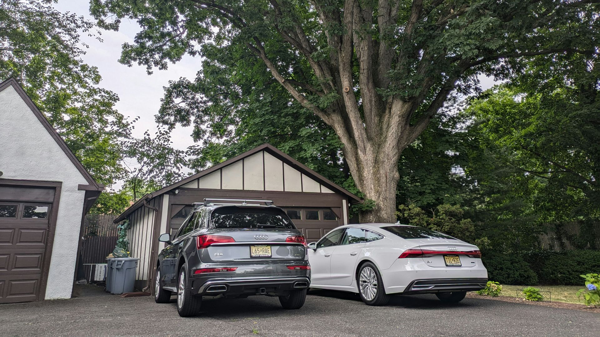 A gray SUV and a white sedan are parked on a gravel driveway in front of a garage under a large, leafy tree.