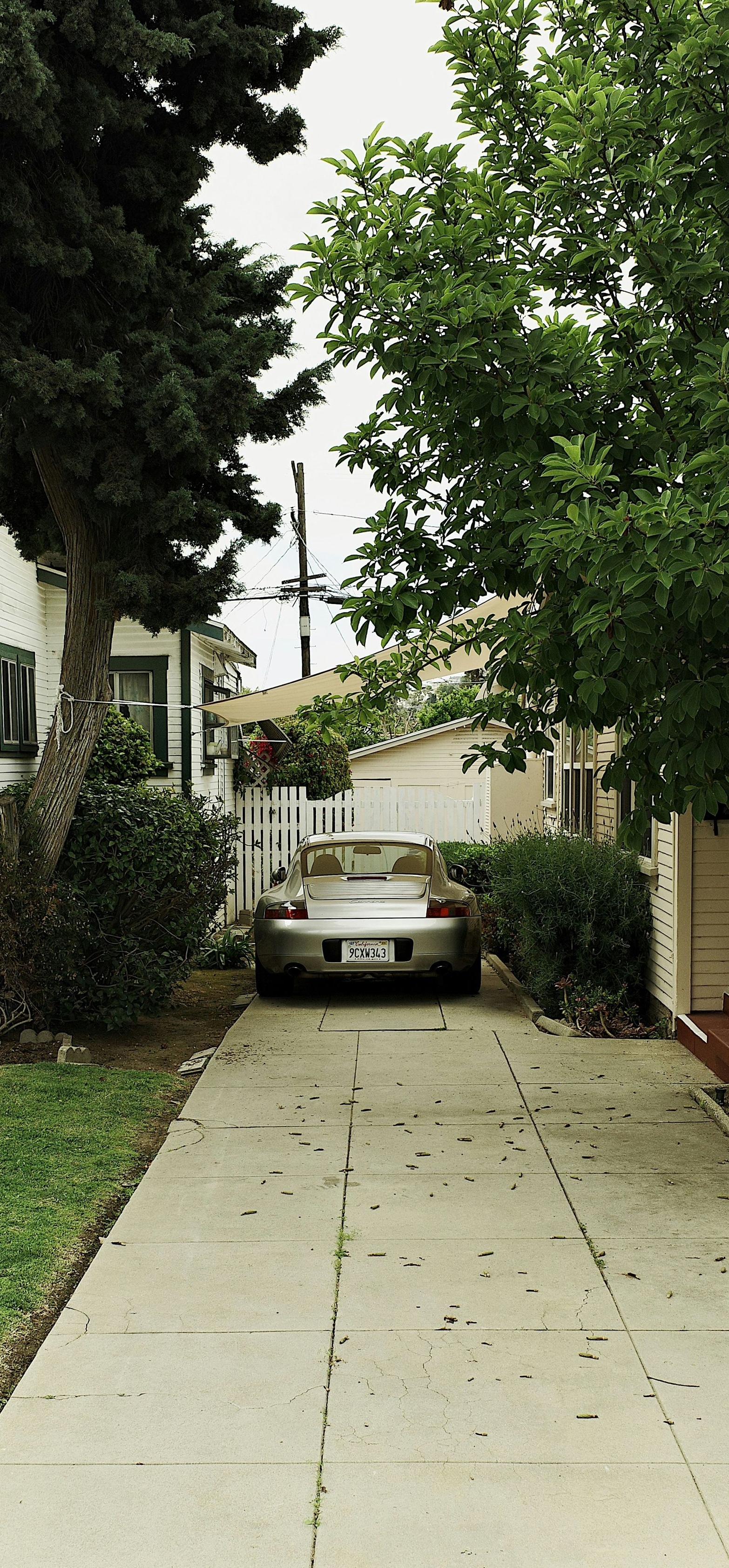 A silver Porsche sports car parked in a narrow driveway between two houses and lush green trees.