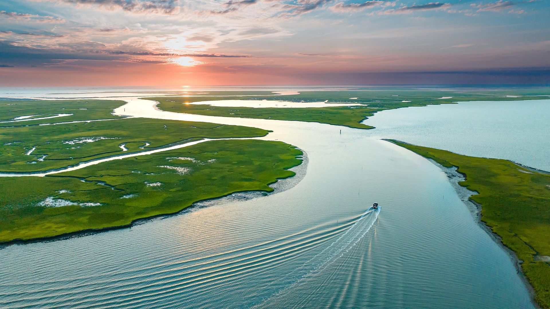 A boat travels on a river through marshland at sunset; orange and pink sky.