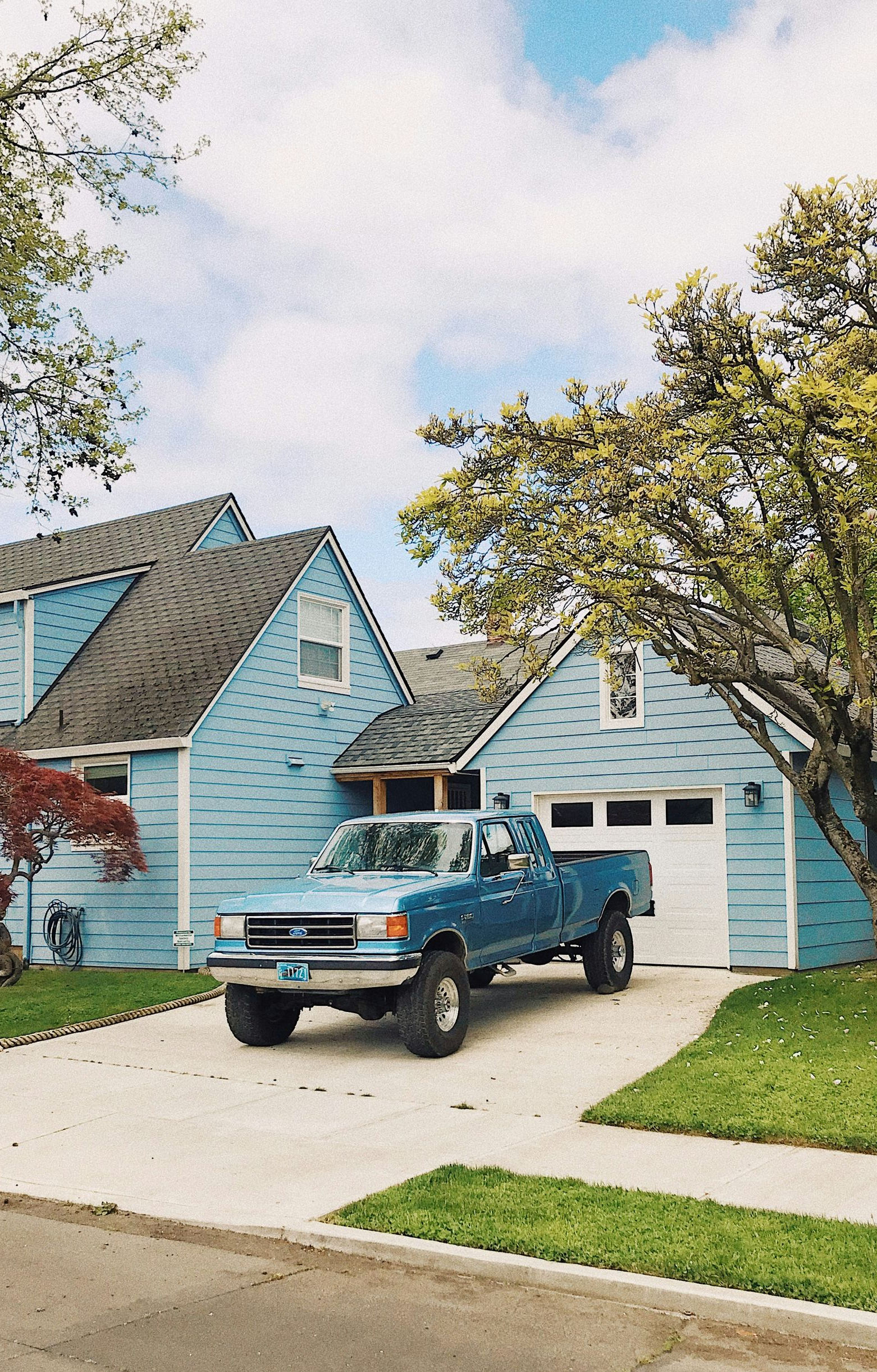 A blue pickup truck parked in the driveway of a blue house with white garage doors and a tree in the yard.