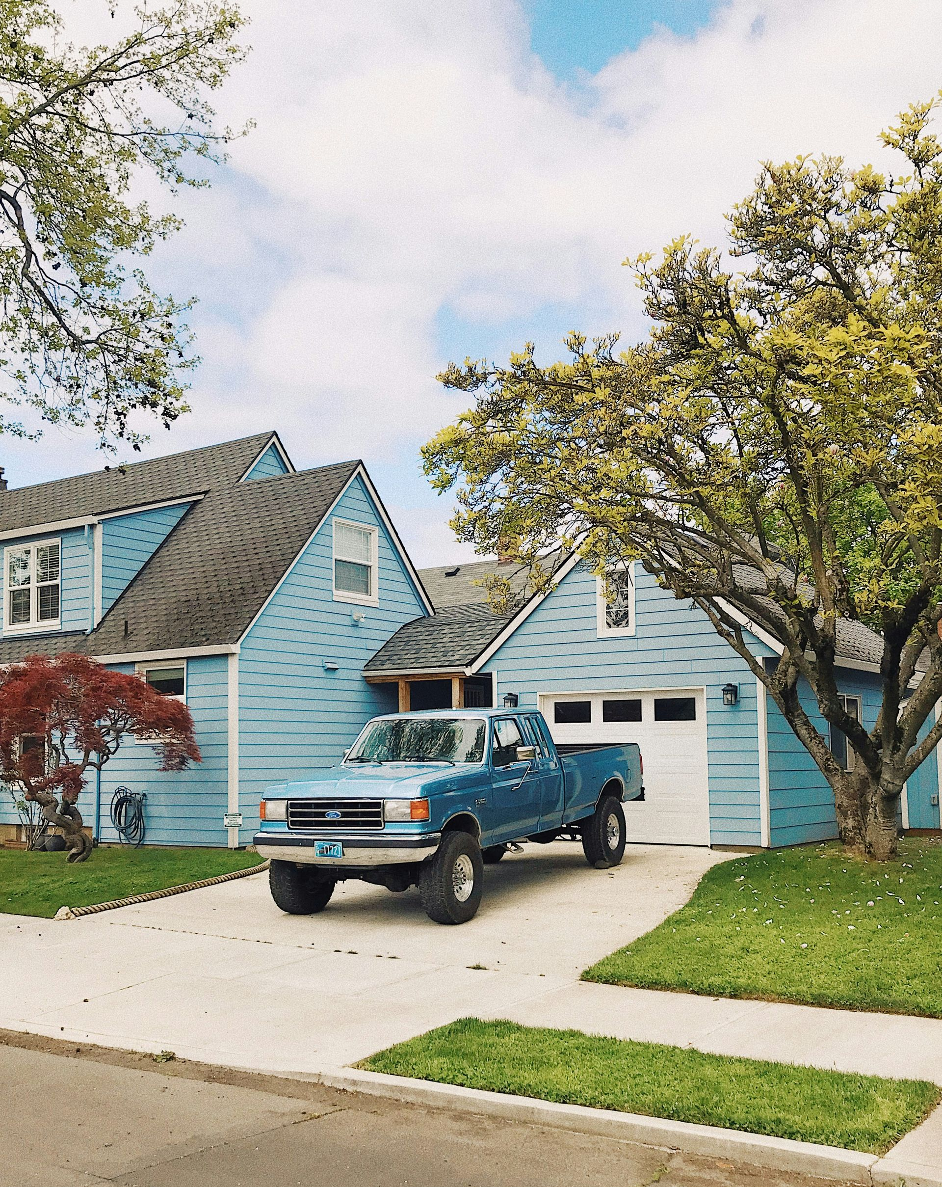 Blue truck parked in driveway of light blue house, green lawn, tree, cloudy sky.