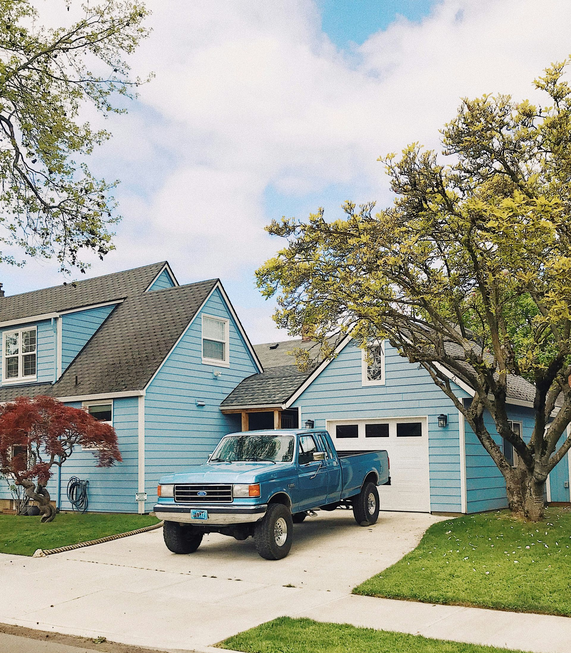 Blue truck parked in driveway of light blue house, green lawn, tree, cloudy sky.