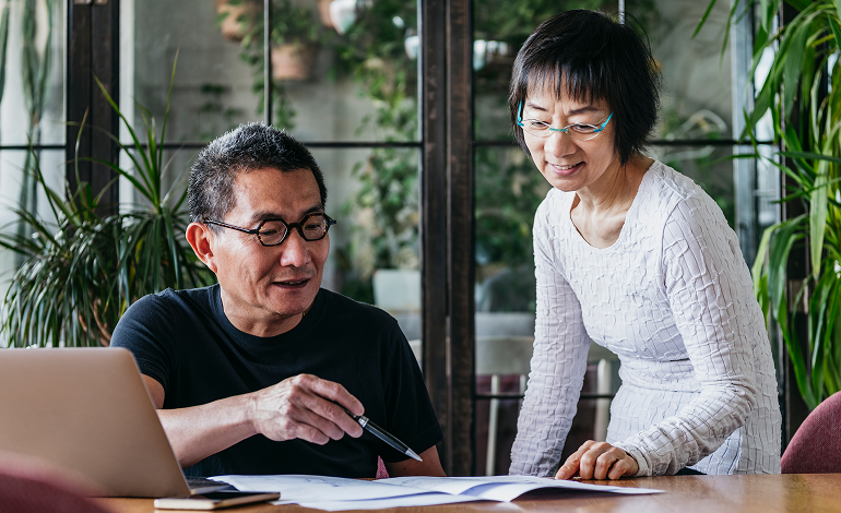 A man and a woman are sitting at a table looking at a laptop.