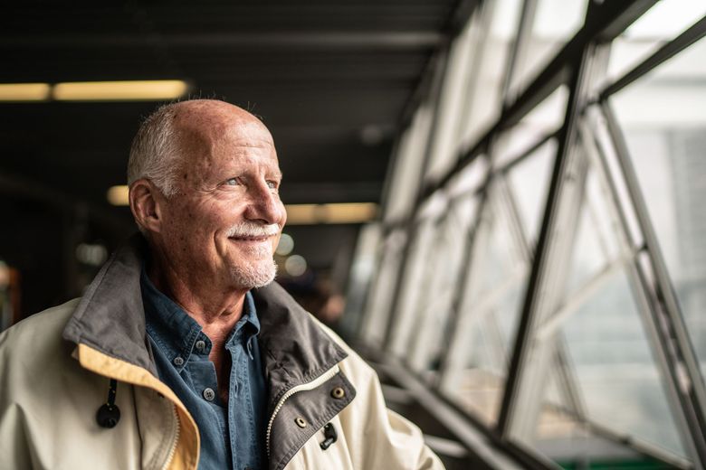 An elderly man is standing in front of a window and smiling.
