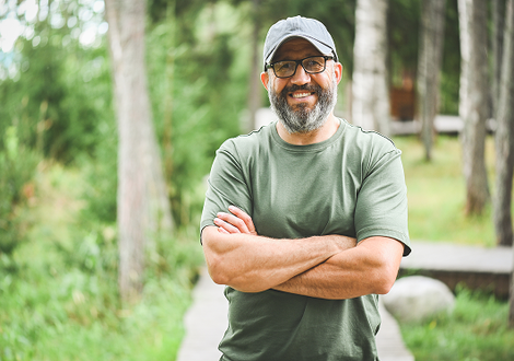 A man with a beard and glasses is standing with his arms crossed in the woods.