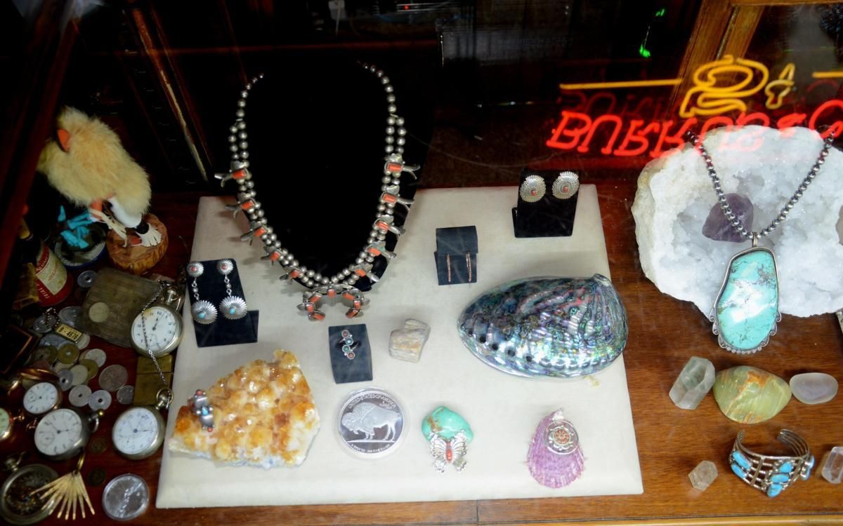 Jewelry display: necklaces, earrings, and pendants with stones and shells on a tan surface. Neon sign in background.