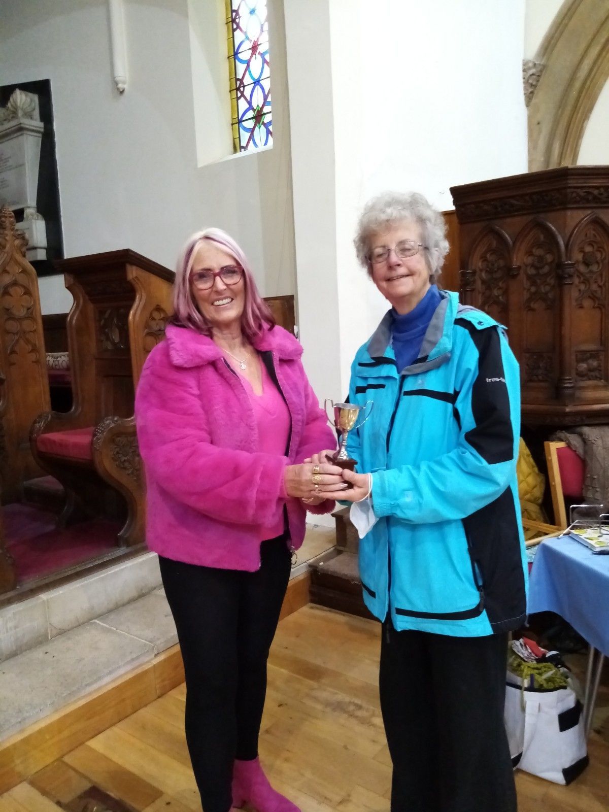 A woman in a pink jacket is receiving a trophy from another woman in a blue jacket