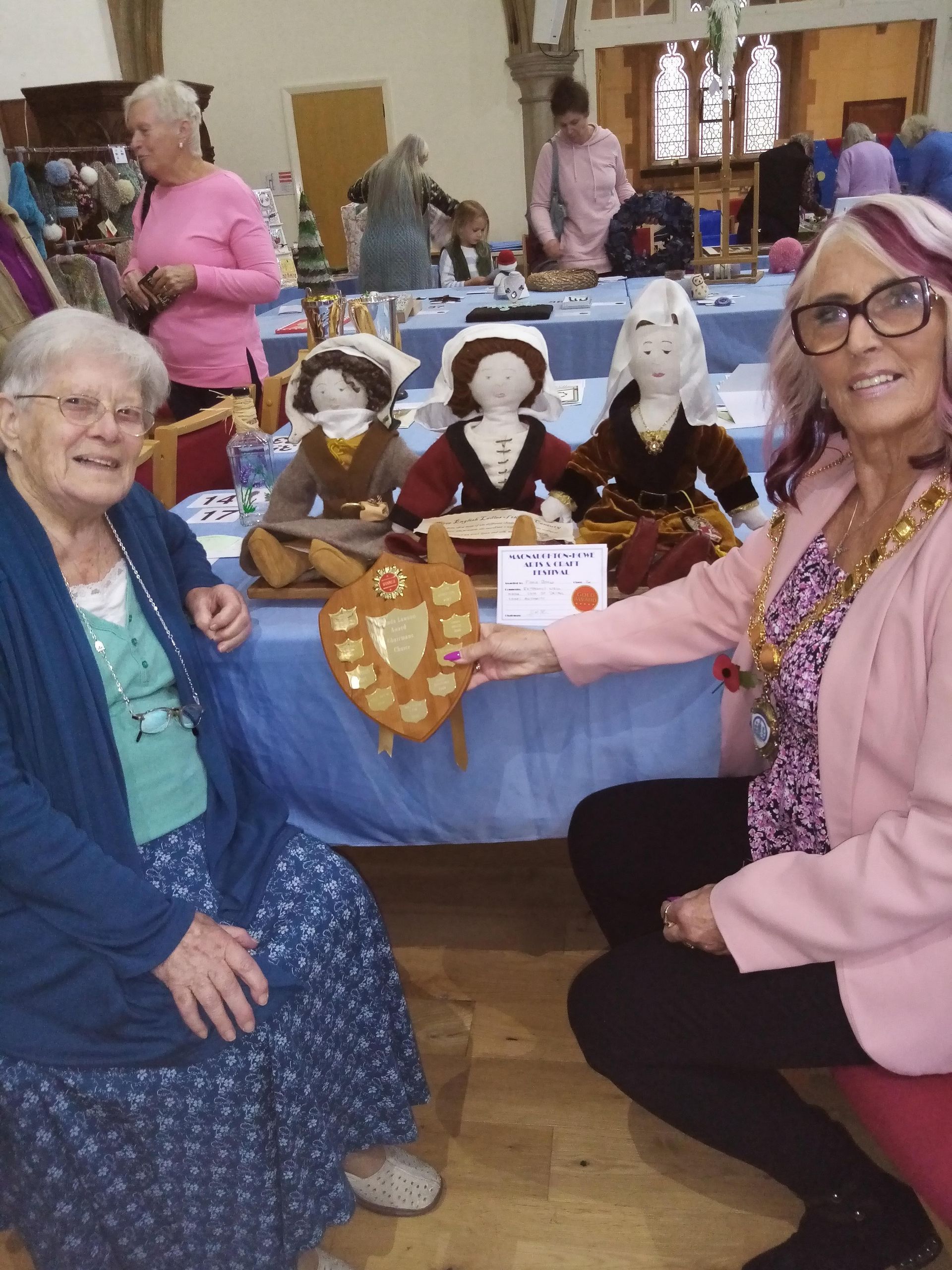 Two women are sitting at a table holding a plaque and dolls