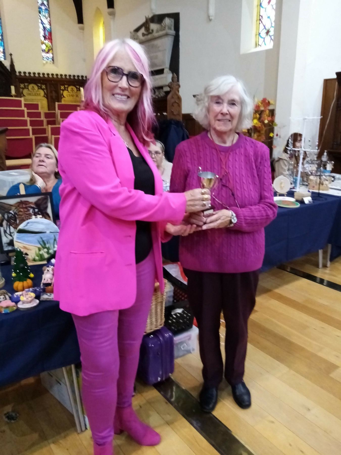 A woman in a pink jacket is handing a trophy to another woman