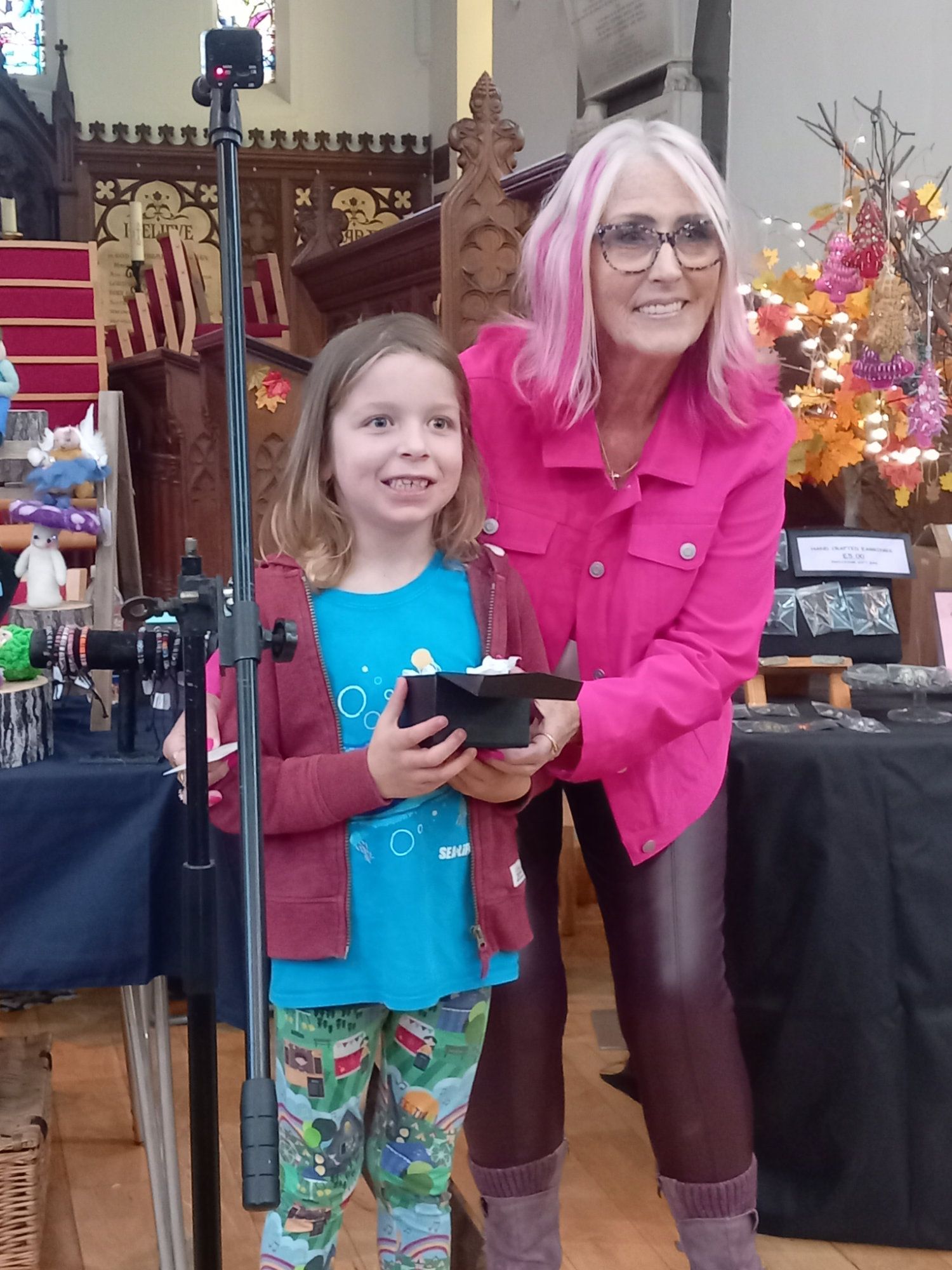 A woman with pink hair is standing next to a little girl holding a box.