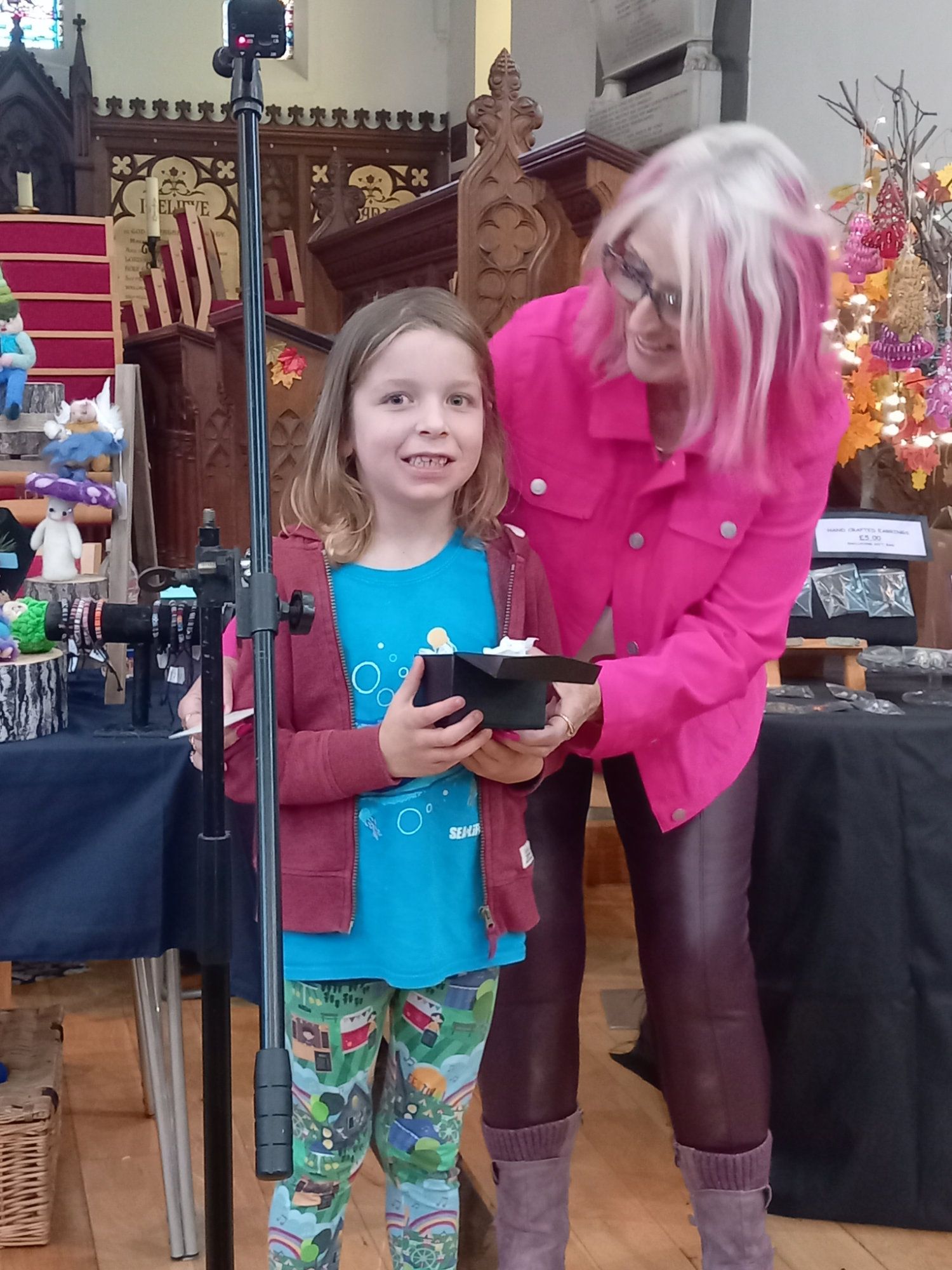 A woman with pink hair is standing next to a little girl holding a box.