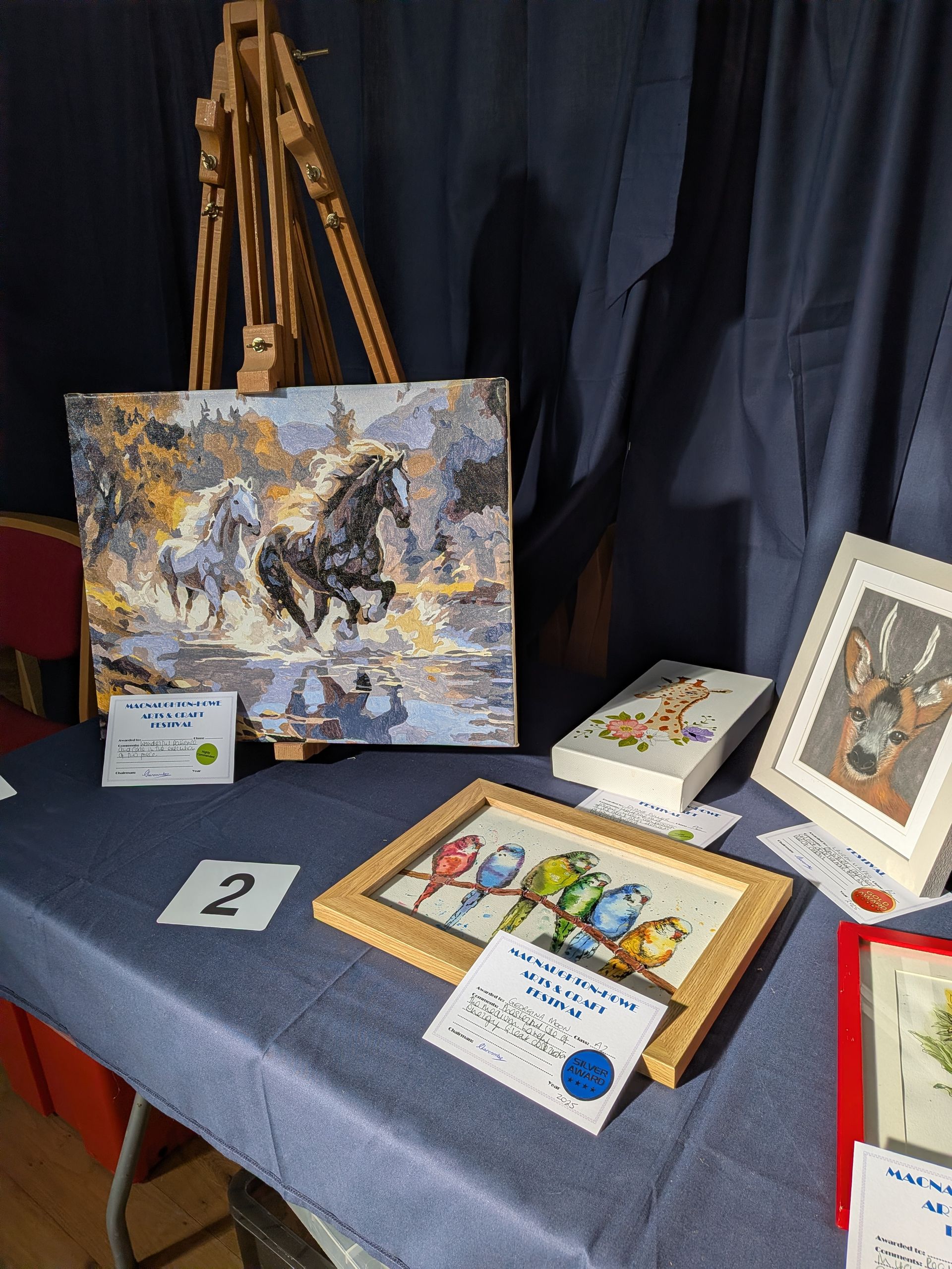 Display of cross-stitch art: horses, birds, and other designs on a blue table. Wooden easel supports a horse scene.