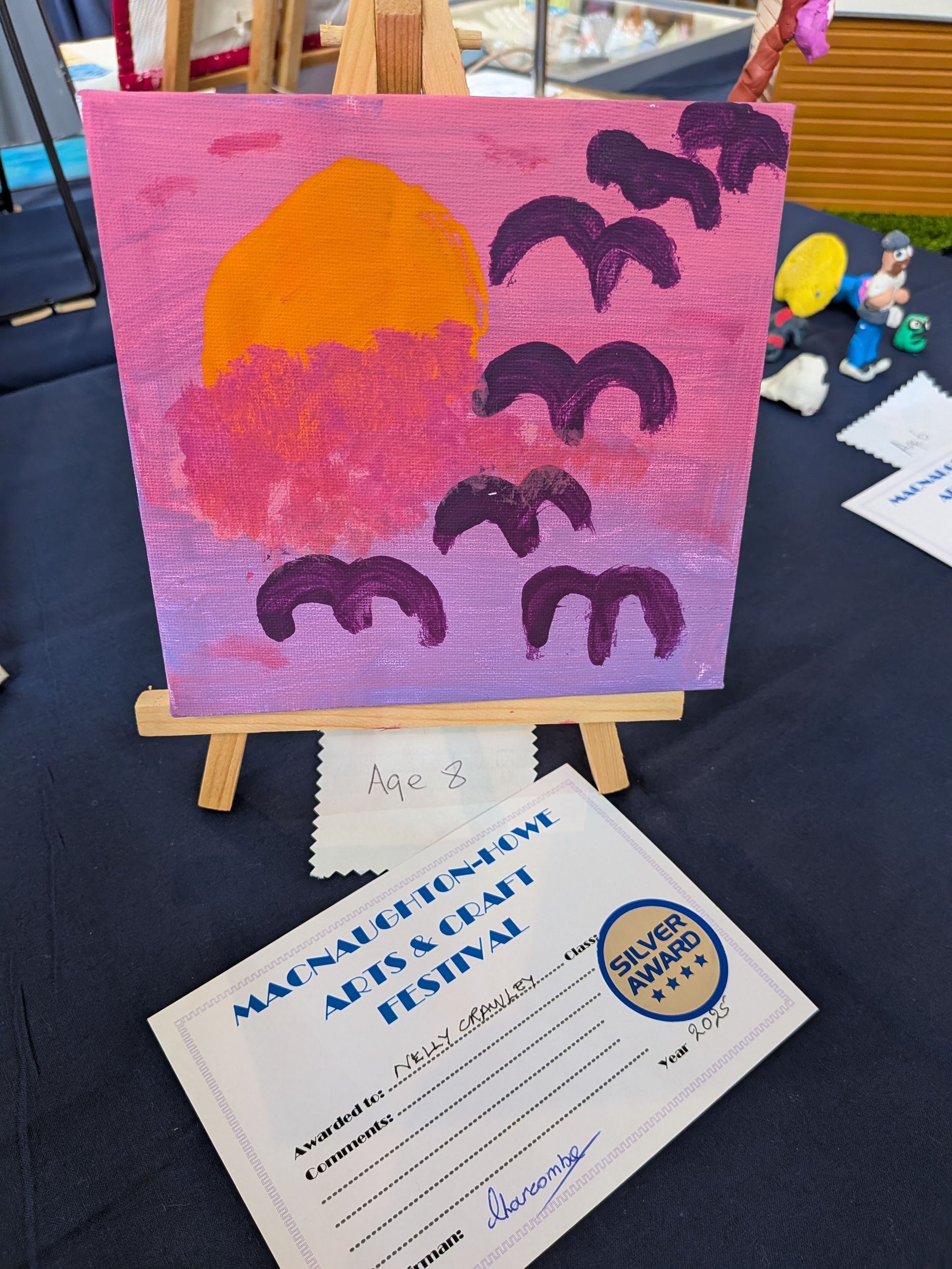 Painting of sunset with birds on a wooden easel, next to a silver award certificate.