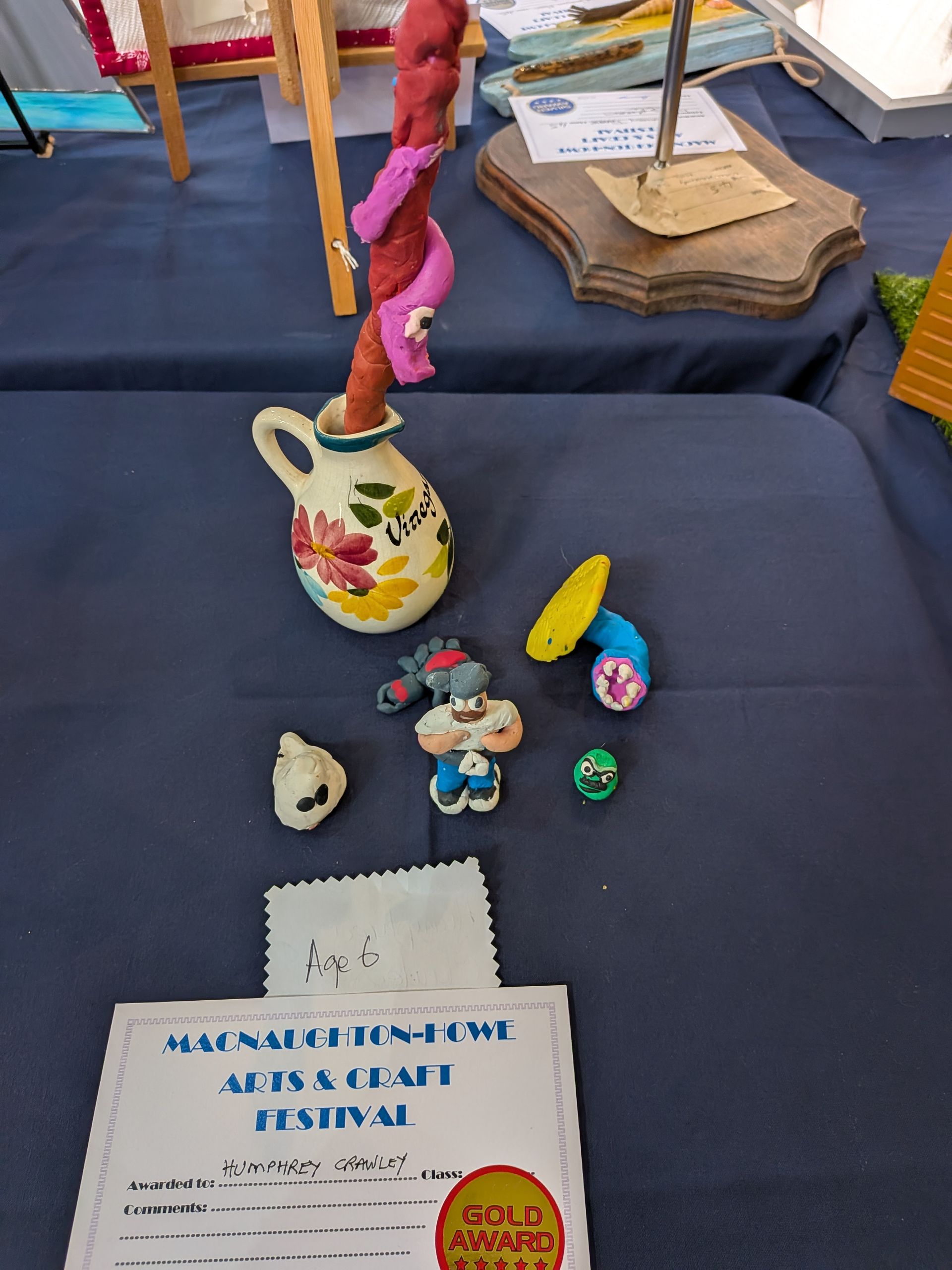 Art display at a craft festival: a vase with a snake sculpture, cartoon figures, and a gold award.
