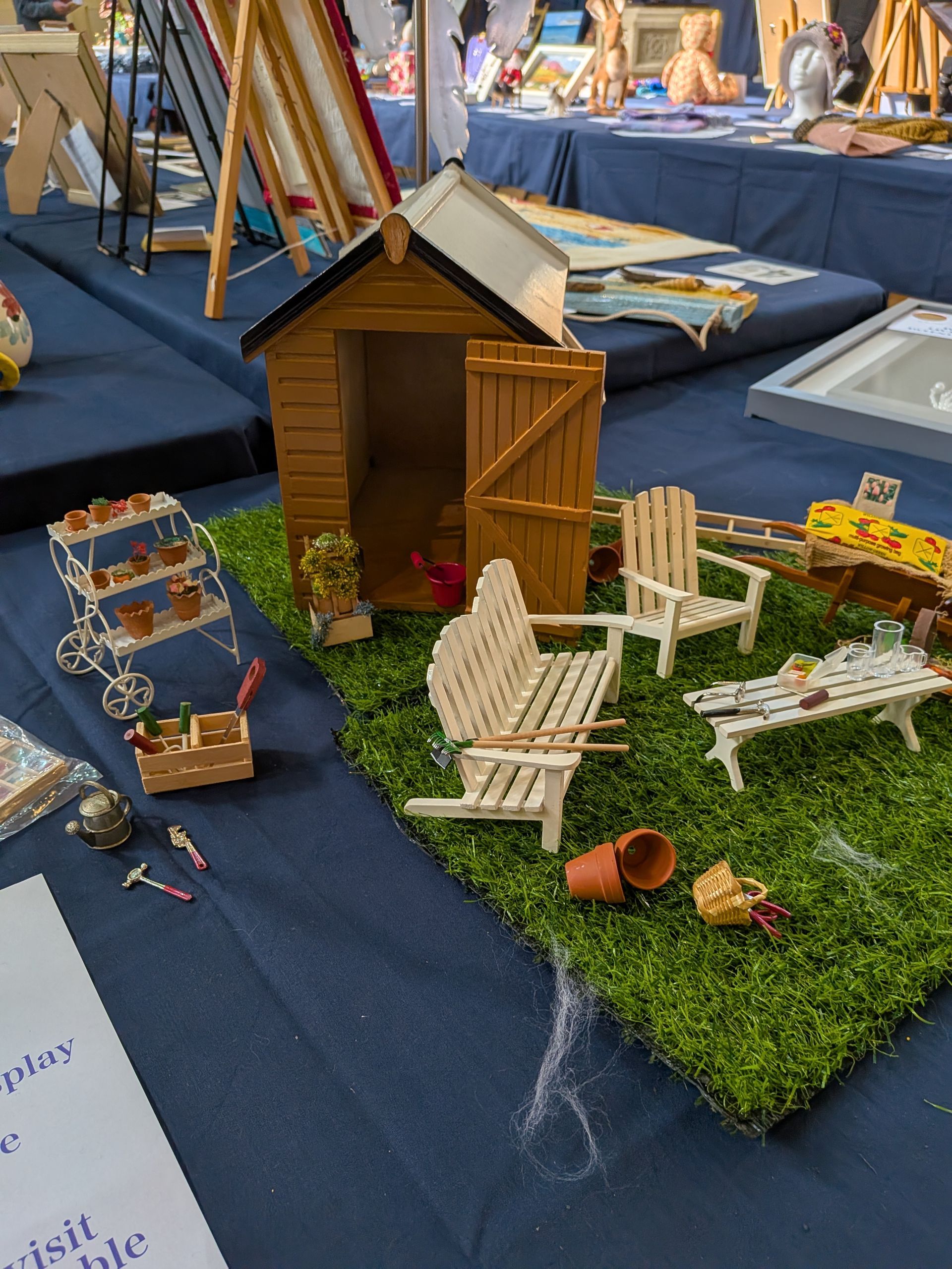 Miniature garden scene with shed, furniture, and grass on a display table.