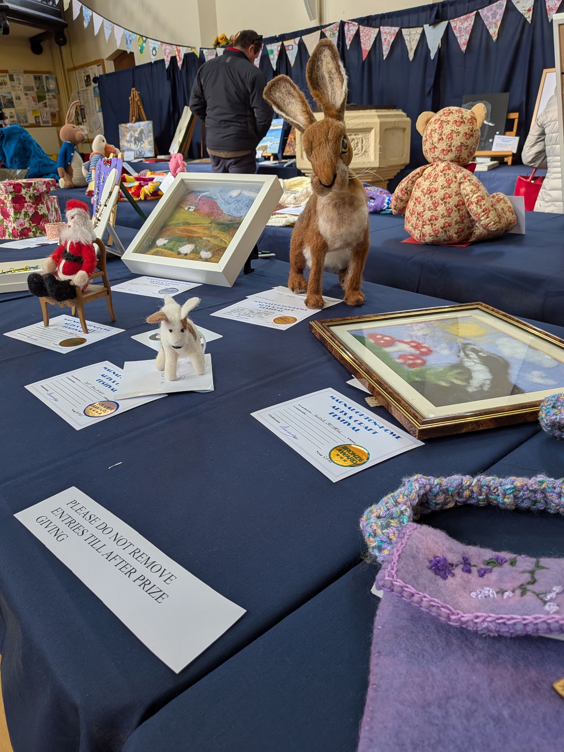 Table of handmade crafts at a fair: toy hare, bear, pictures, Santa, and other items.