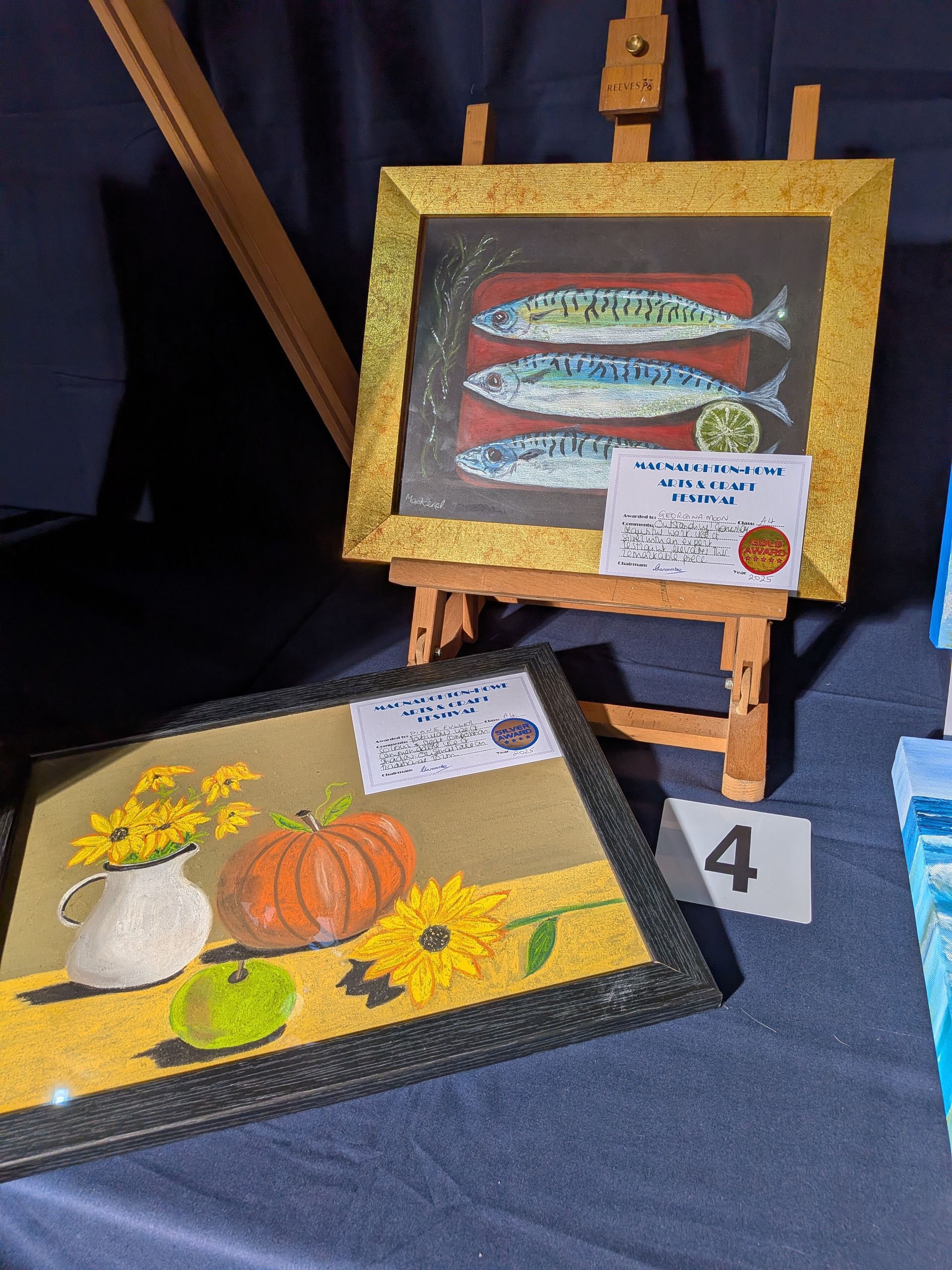 Two still-life paintings: fish and a floral/fruit arrangement, on easels, on a dark blue surface.