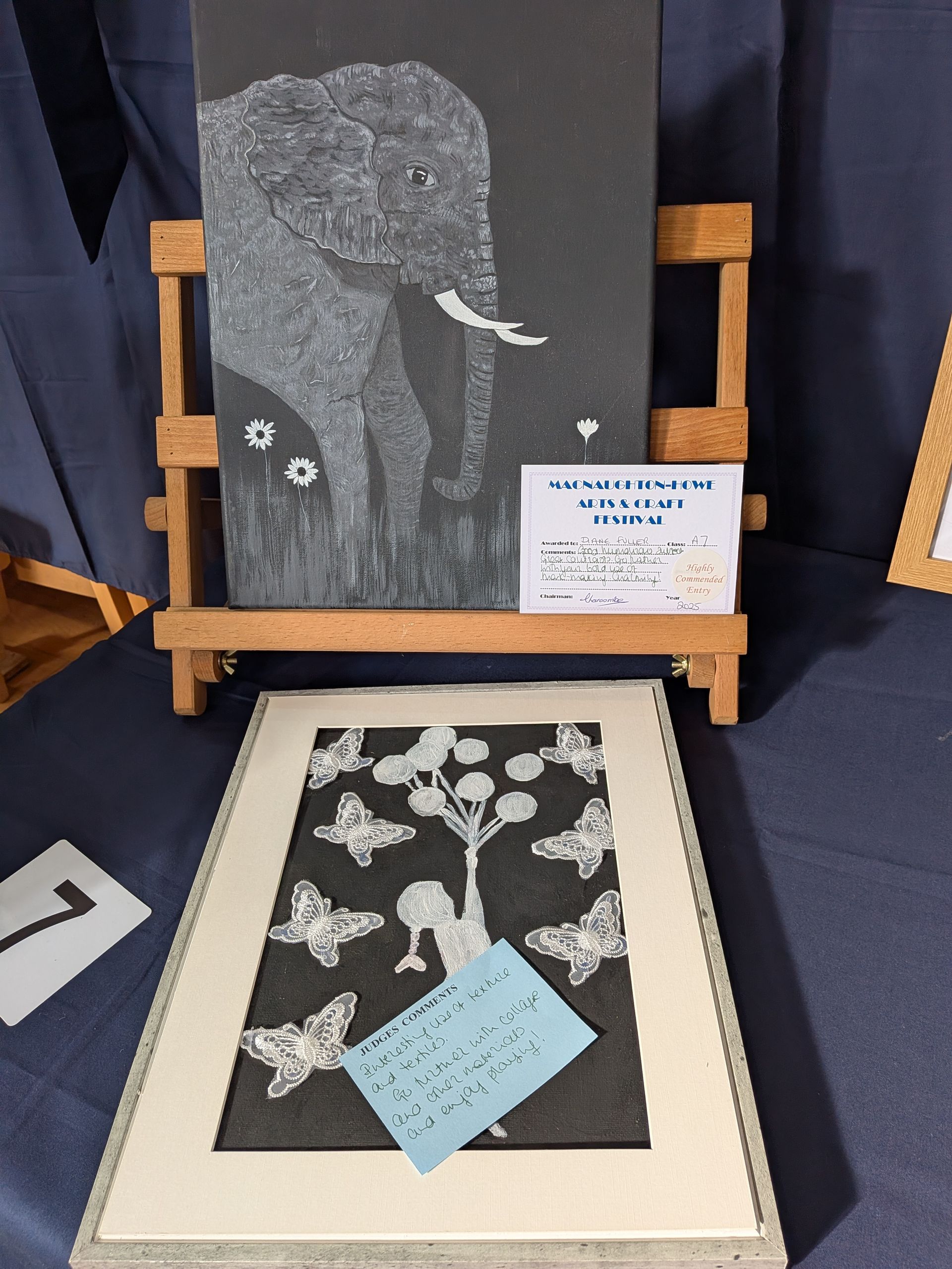 Art display: elephant etched on black, framed butterfly scene, on small wooden easels; blue tablecloth.