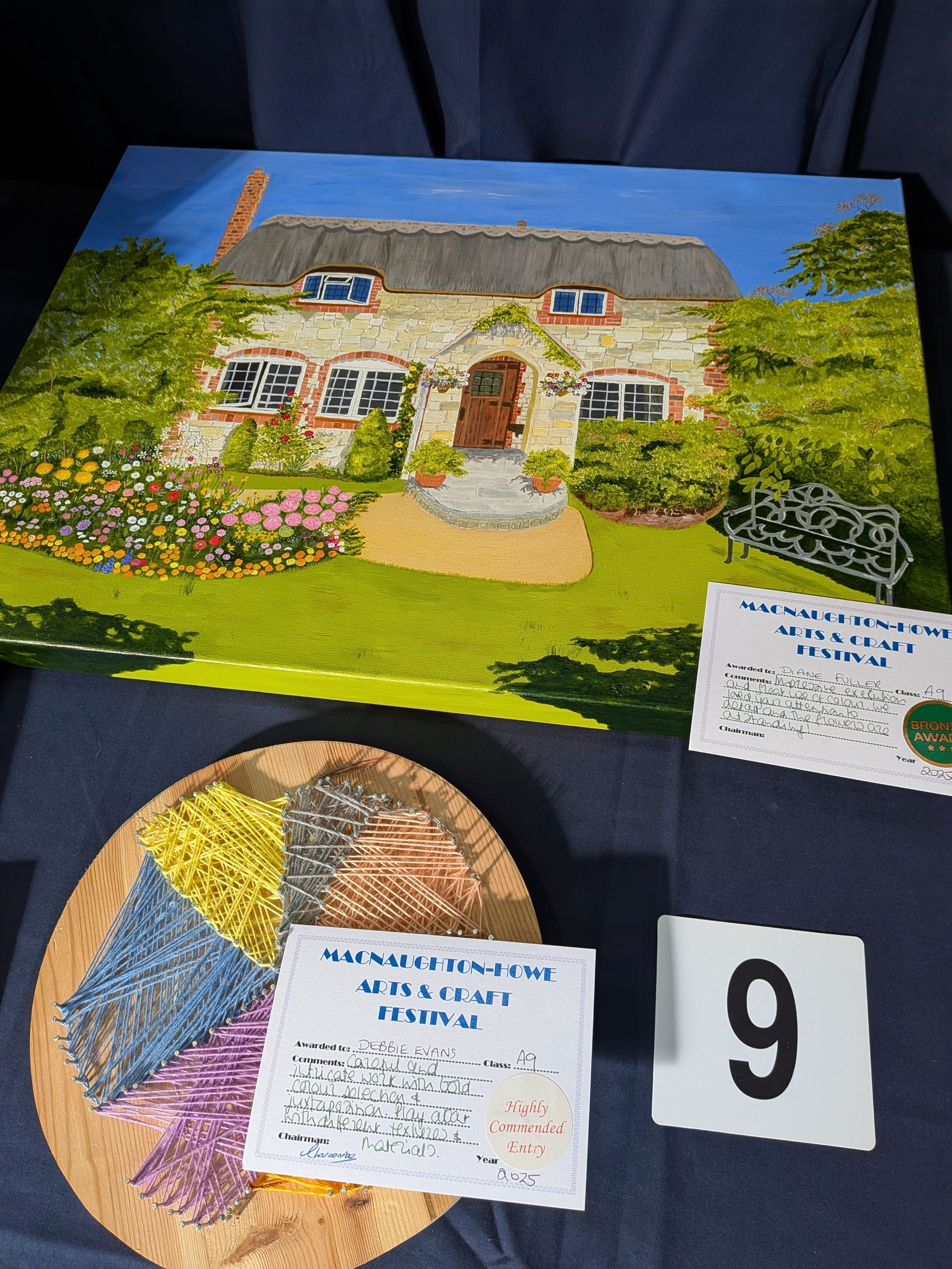Painting of a cottage with sample yarns, a number 9, and a description card on a blue surface.