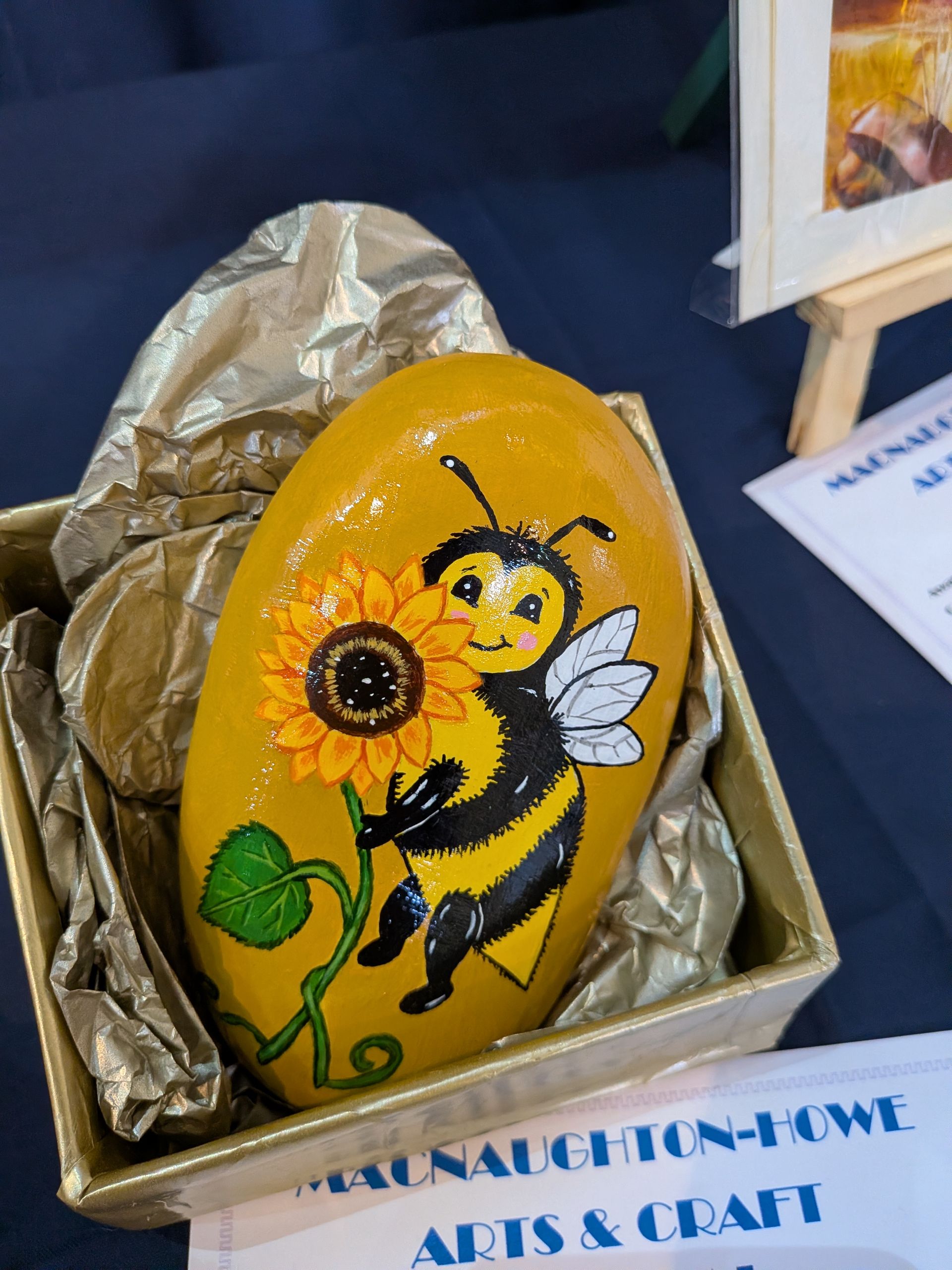 Painted rock with a smiling bee holding a sunflower, displayed in a golden box with gold tissue paper.