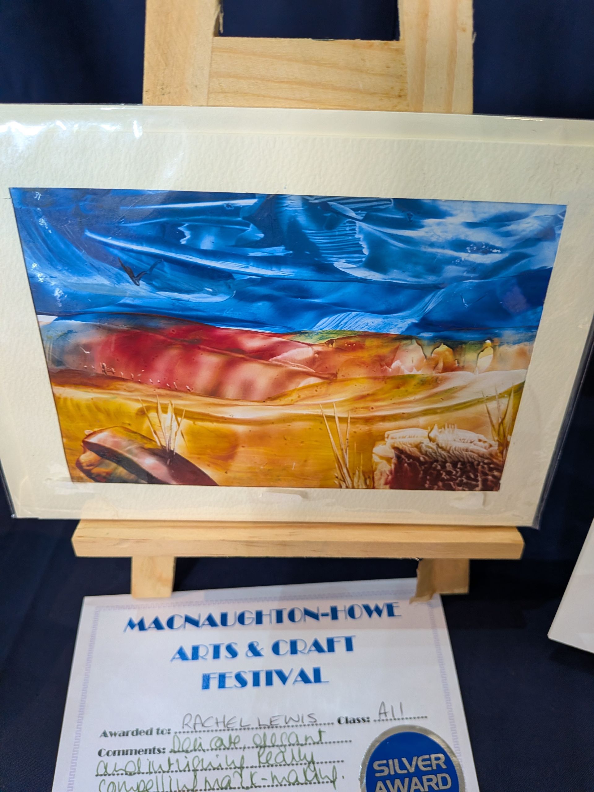 Watercolor landscape painting on easel; blue sky, red and yellow land. Silver award certificate.