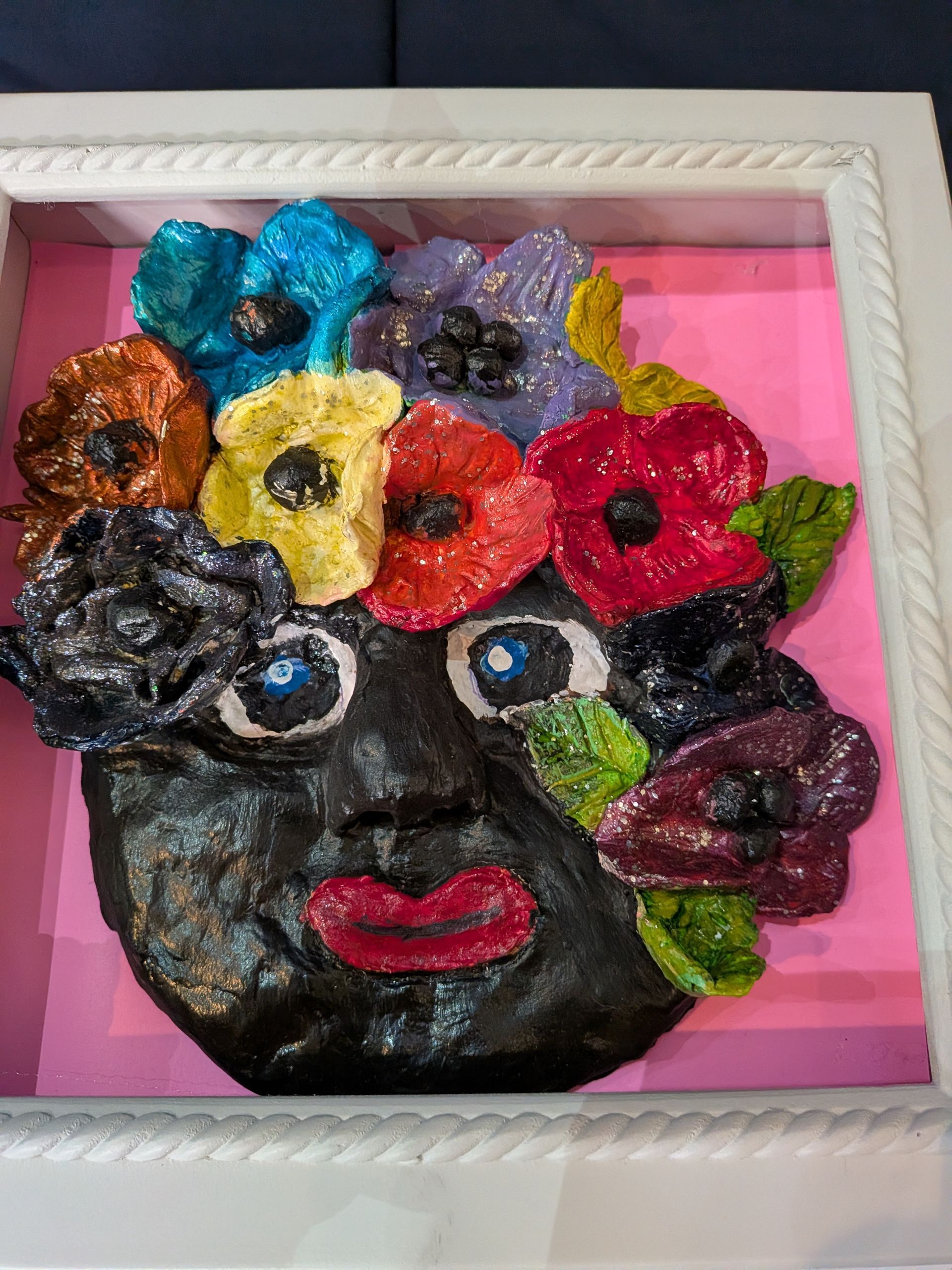 Sculpted face with flowers, in a white shadow box. Black face, colorful flowers, pink background.