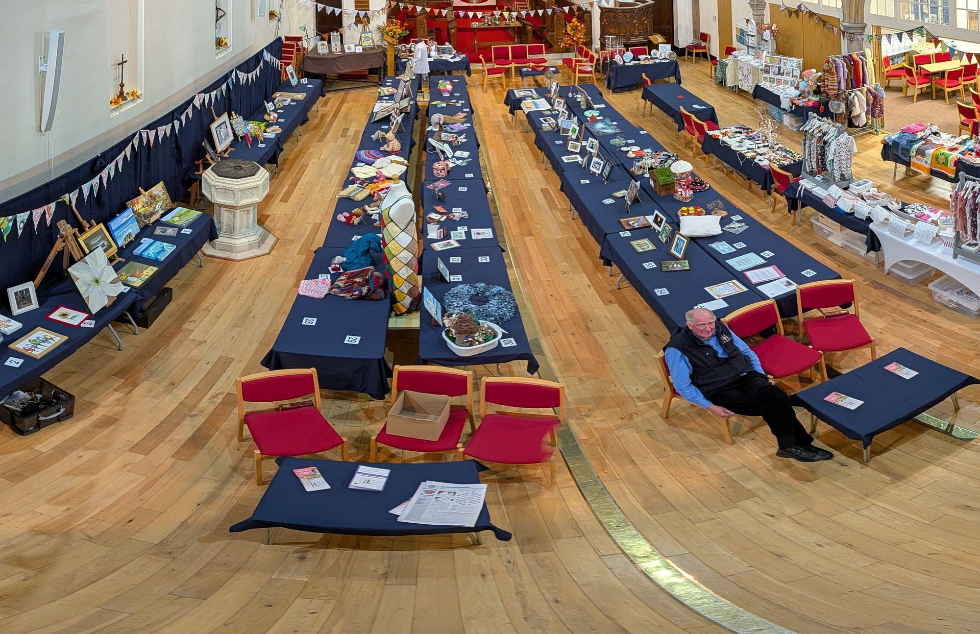 A craft fair in a hall with long tables displaying goods. A man sits by a small table.